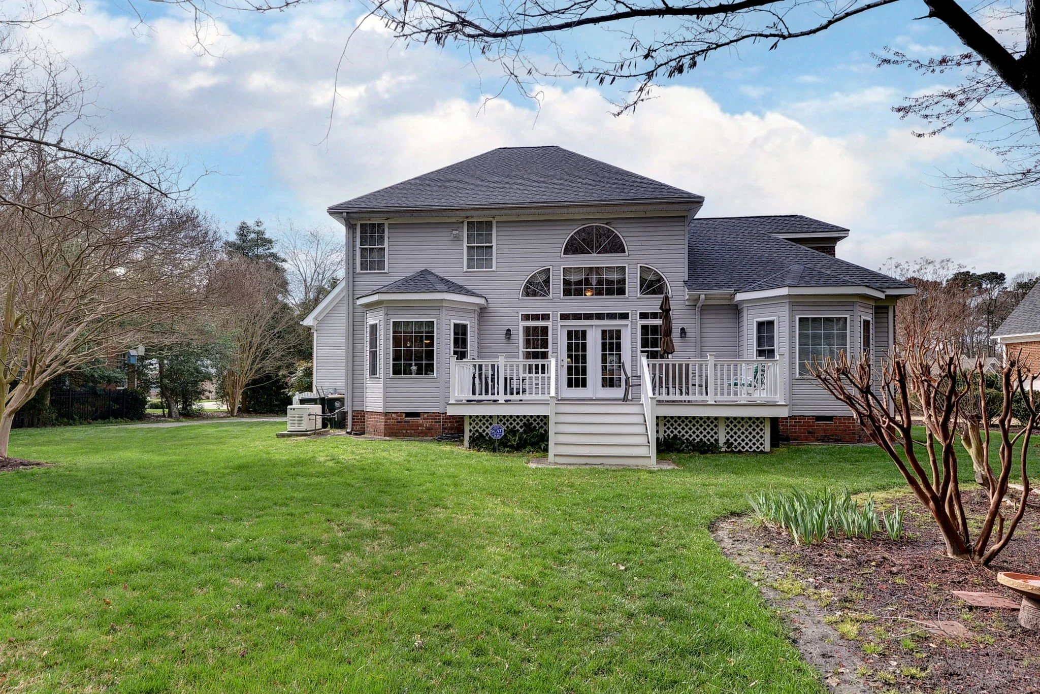 The back of a two-story house with gray siding, a black roof, and a white deck with stairs leading to a green lawn. The house has large windows and a glass door, with trees and a cloudy sky in the background.