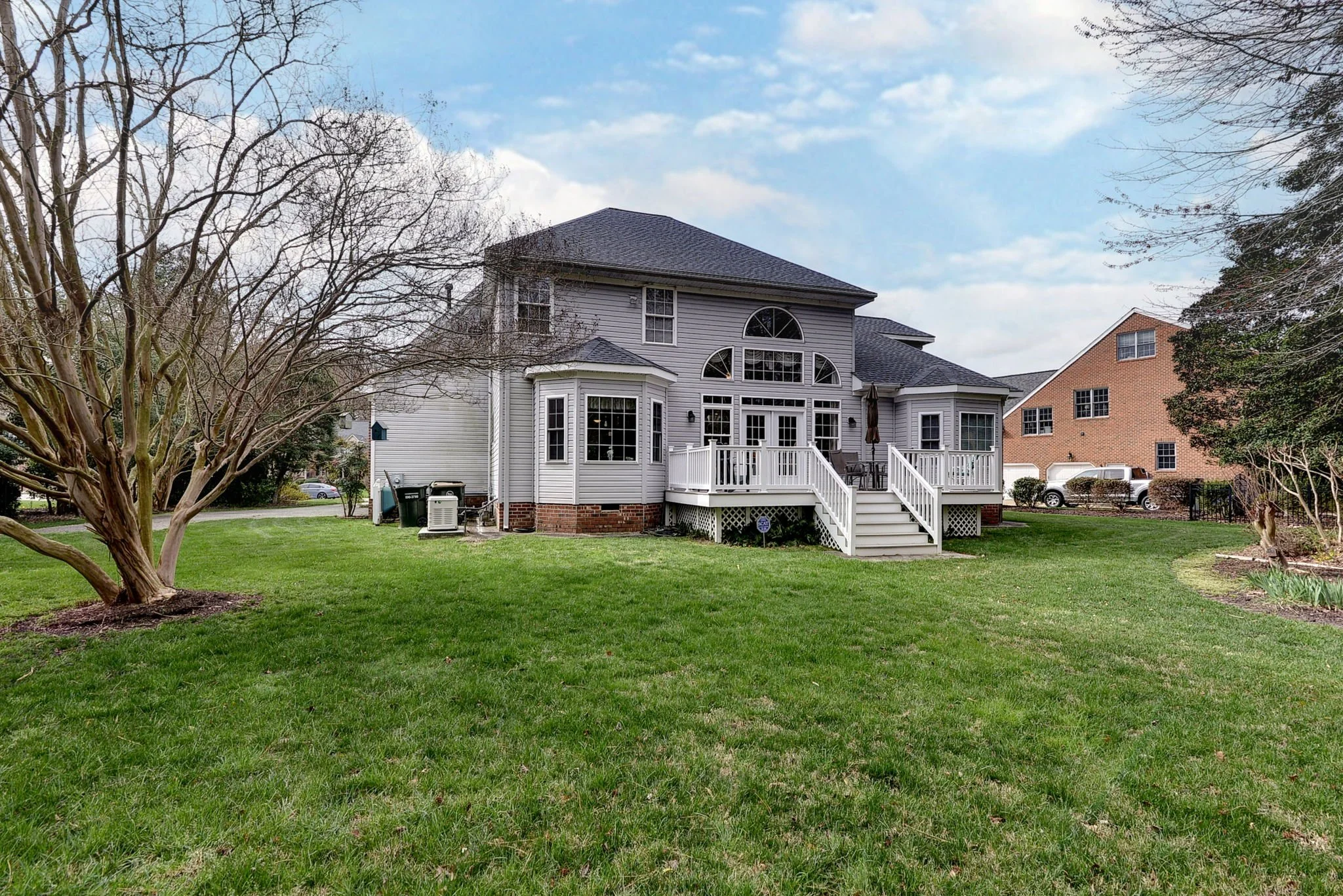 Large gray house with a backyard deck, situated on a well-maintained grassy lawn, with neighboring house visible in the background and a partly cloudy sky overhead.