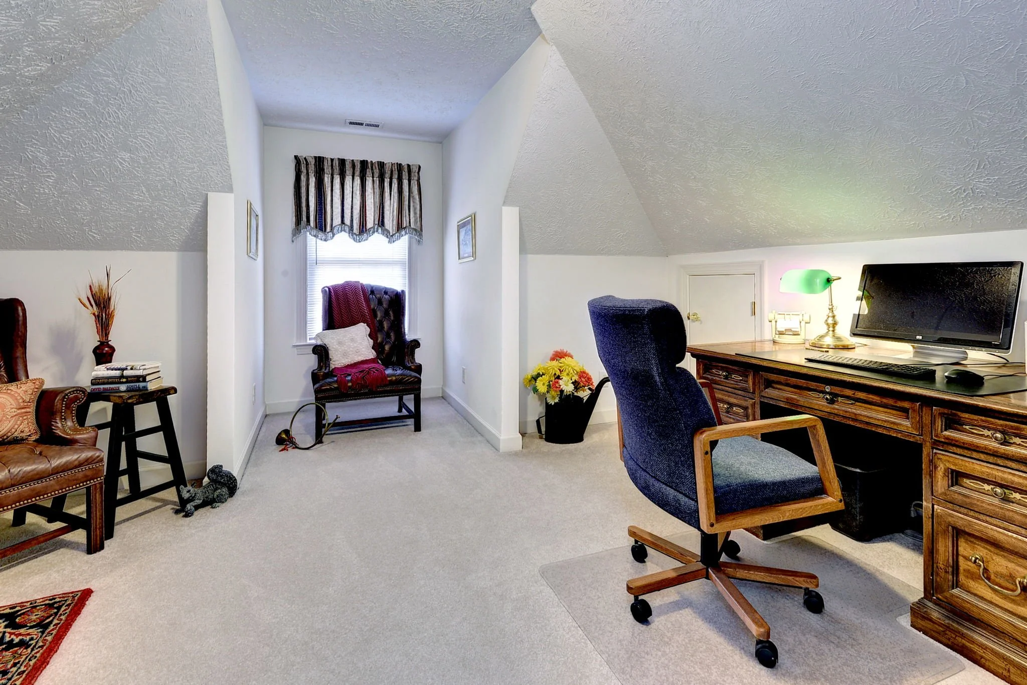 A cozy home office space with a wooden desk, a computer monitor, a green desk lamp, a blue office chair, a black plant pot with yellow flowers, and a small window with curtains. There is a plush armchair with throw pillows and a blanket, and a side t