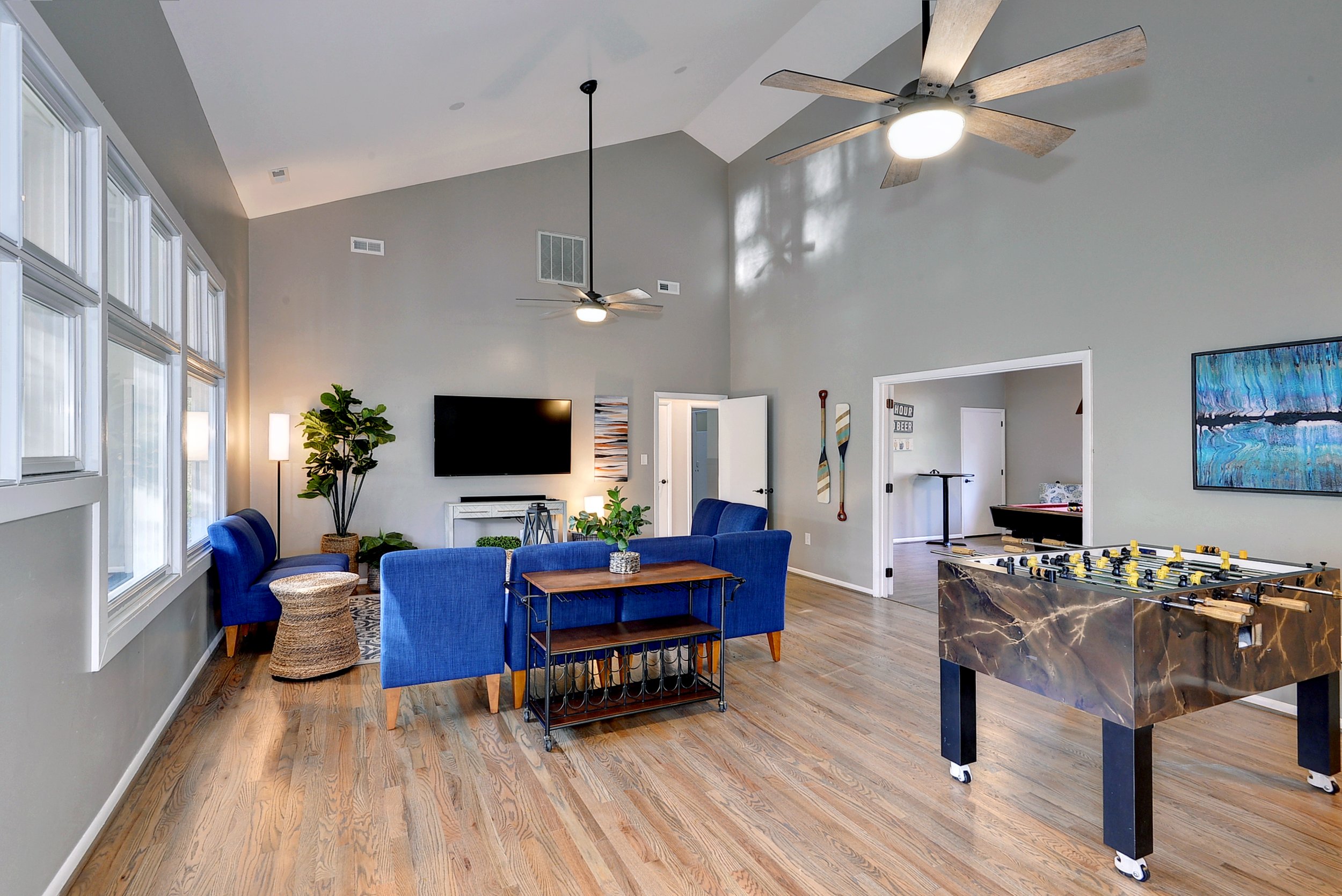 Spacious living room with gray walls, large windows, hardwood floors, a blue sofa and chairs, a wall-mounted TV, ceiling fans, a foosball table, and wall art.