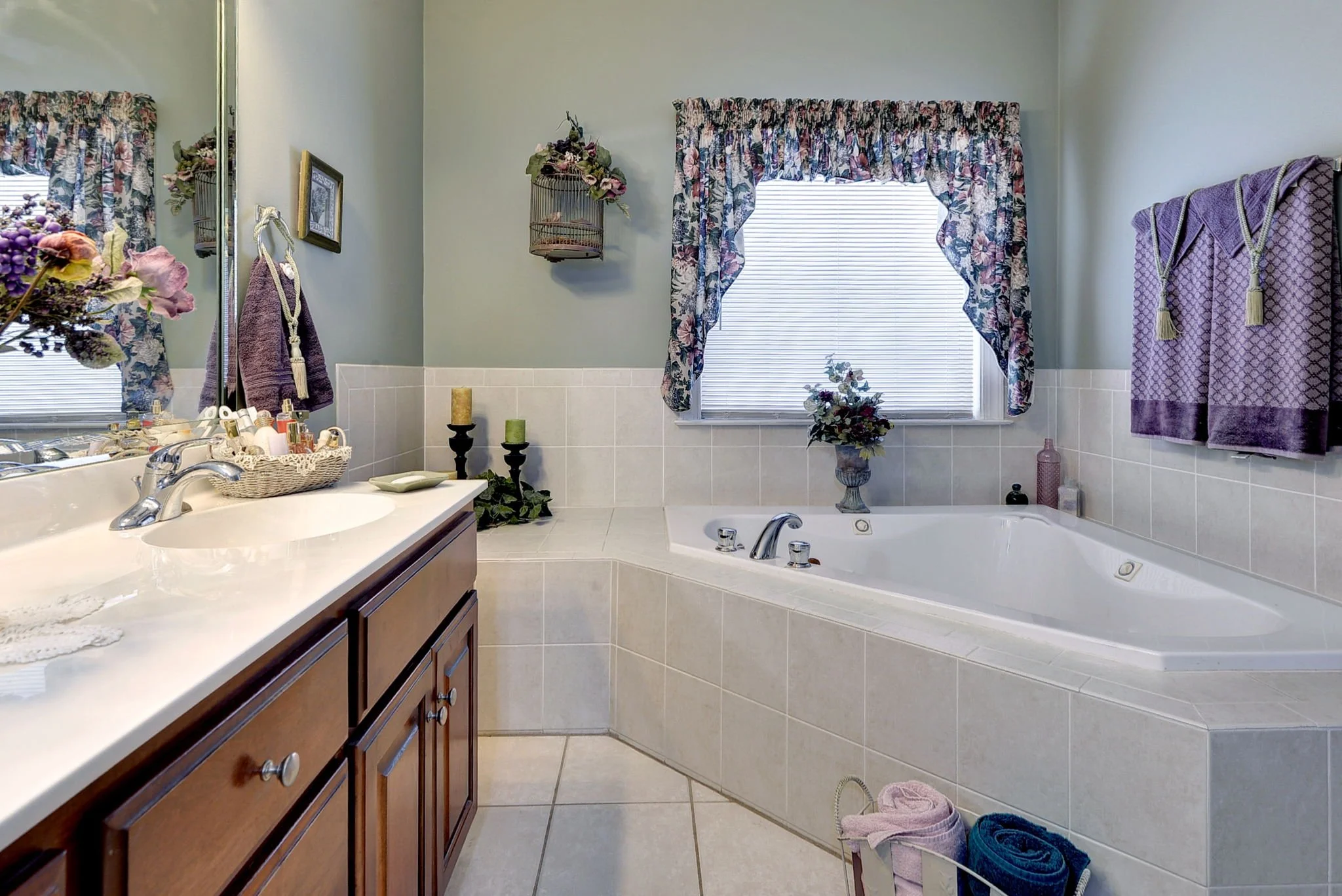 A bathroom with a white countertop, brown cabinets, and a large mirror. The room has a corner bathtub beneath a window with floral curtains. Decor includes candles, flower arrangements, and purple towels.