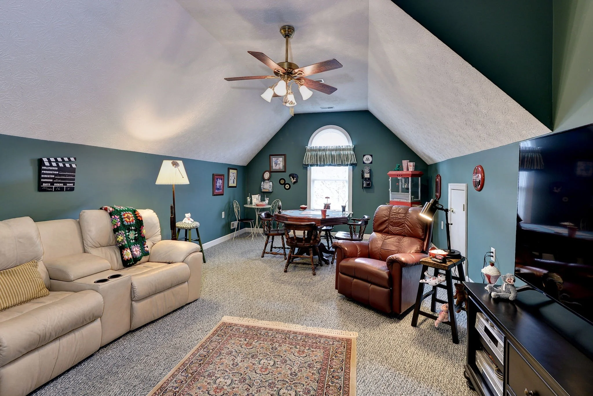 Living room with beige sofa, leather recliner, small side table with lamp, dining table with chairs, TV, and decorative items, under a vaulted ceiling with ceiling fan, blue-green walls, and a window with curtains.