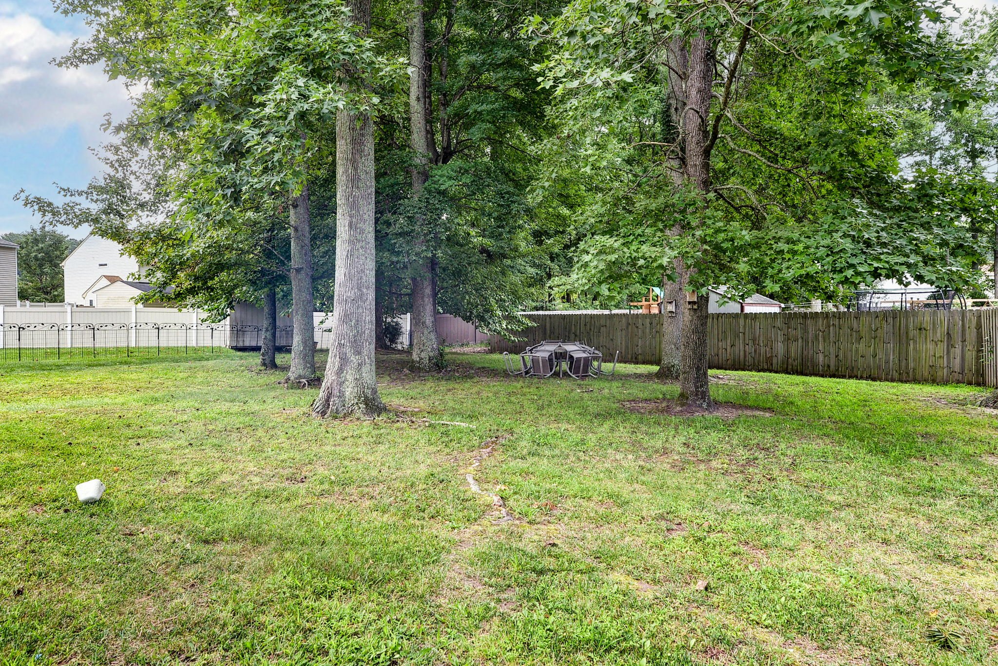 Backyard with green grass, tall trees, a wooden fence, an outdoor table with chairs, and neighboring houses in the background.