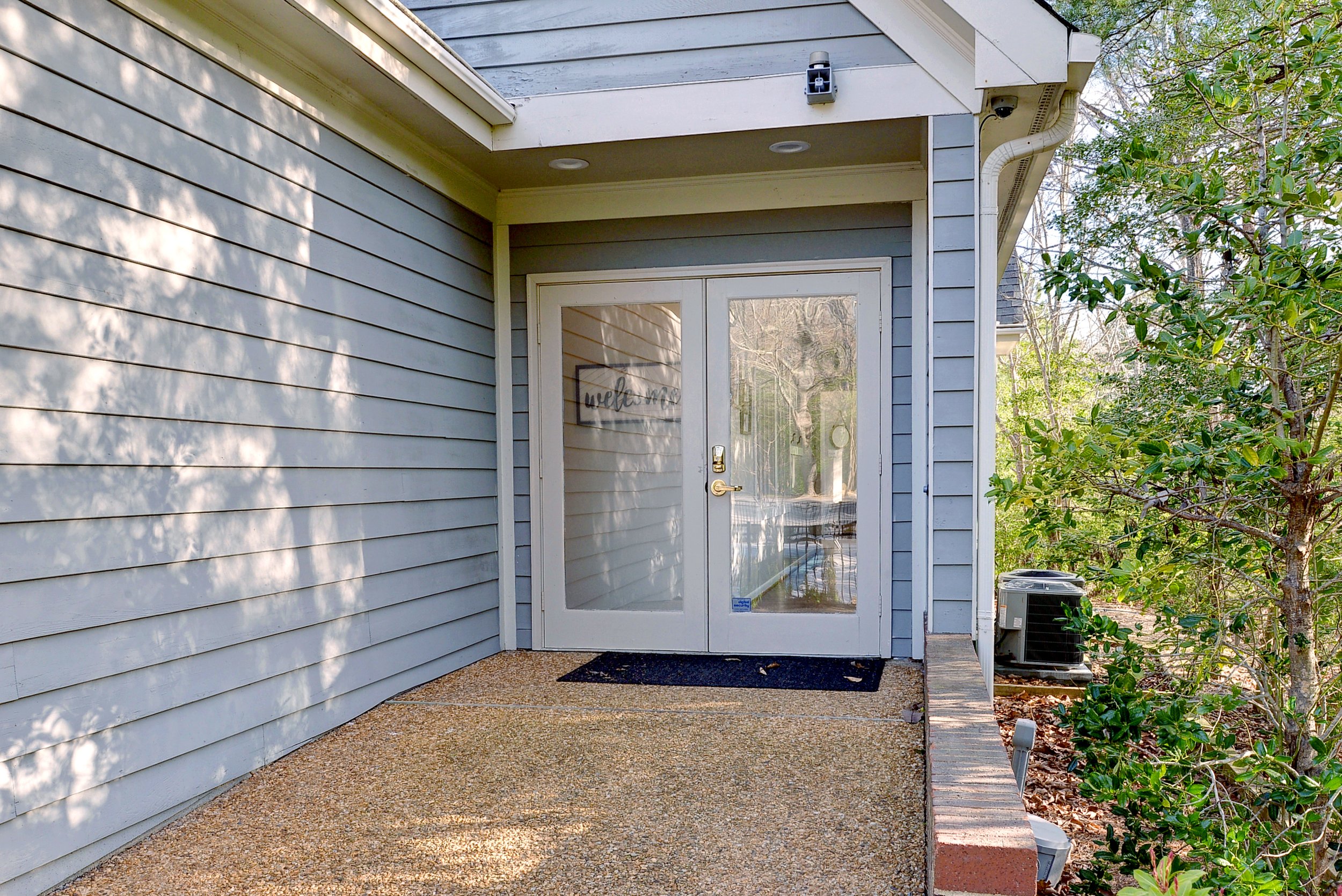 Exterior rear door of a house with glass double doors, surrounded by blue siding, with an outdoor air conditioning unit and greenery nearby.