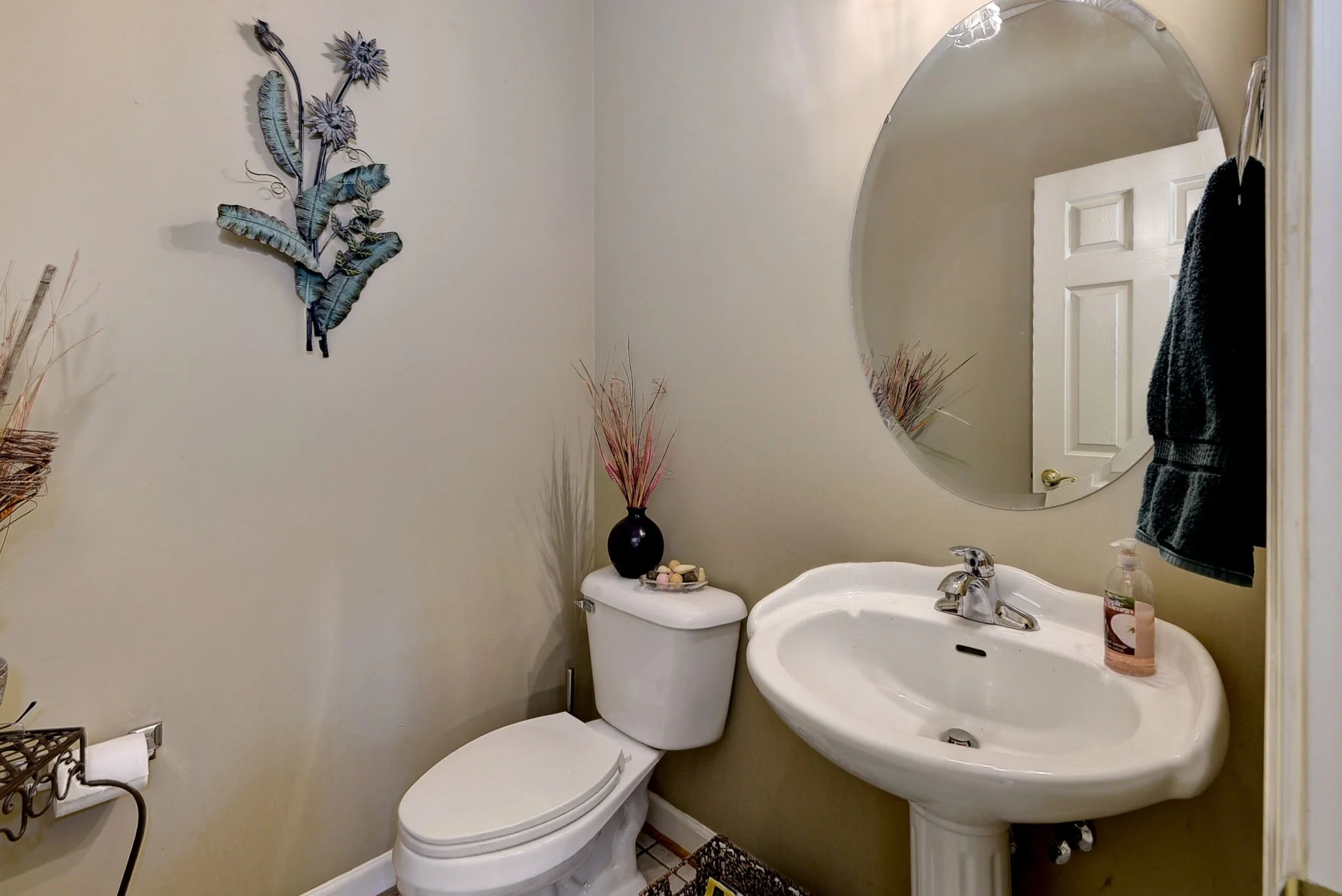 A small bathroom with a white toilet, a white pedestal sink, a large round mirror, a black hanging towel, a black vase with dried flowers on the toilet, and decorative wall art of metal flowers and leaves.