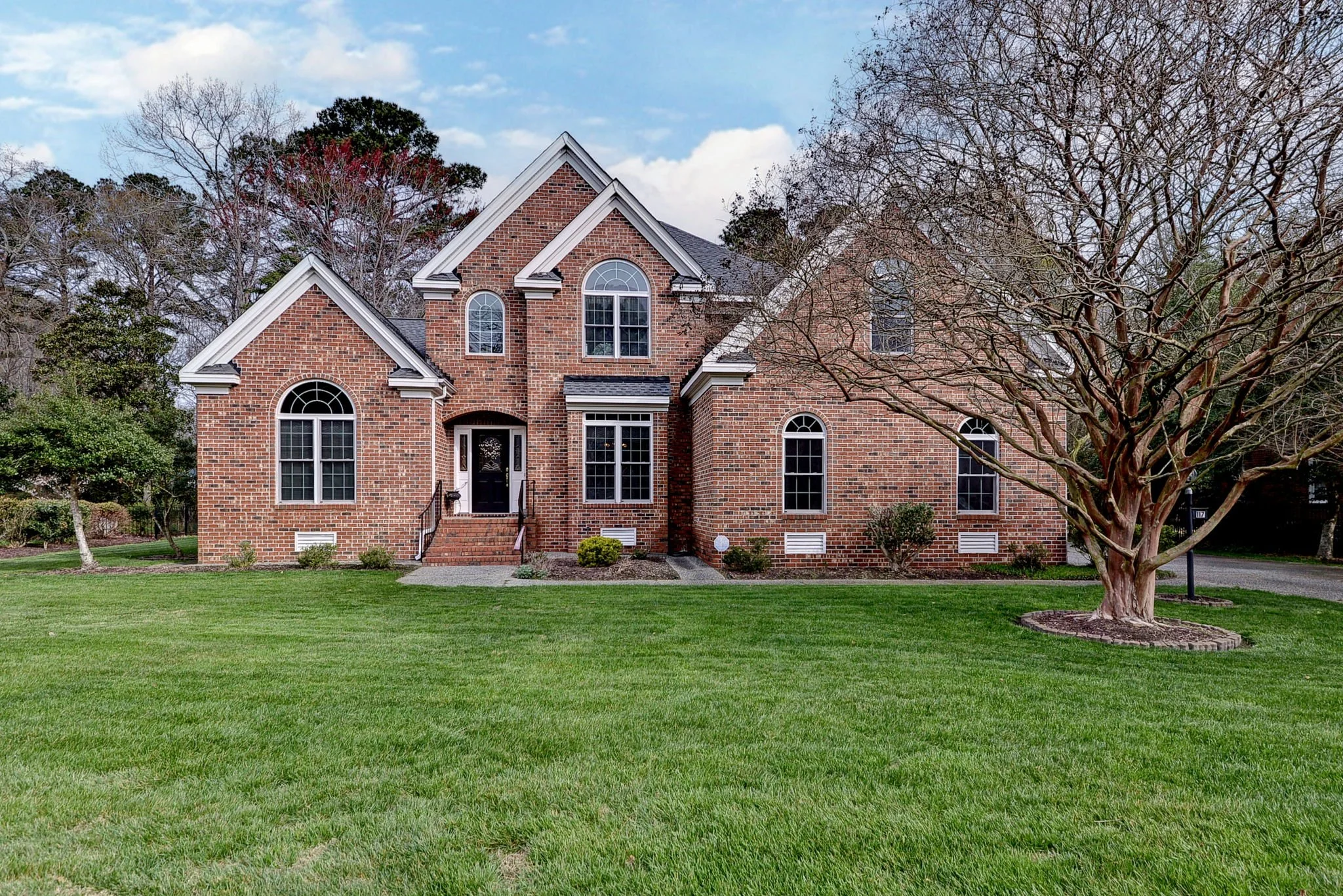 Front view of a large red brick house with multiple gables and arched windows, surrounded by a well-maintained green lawn and a leafless tree.