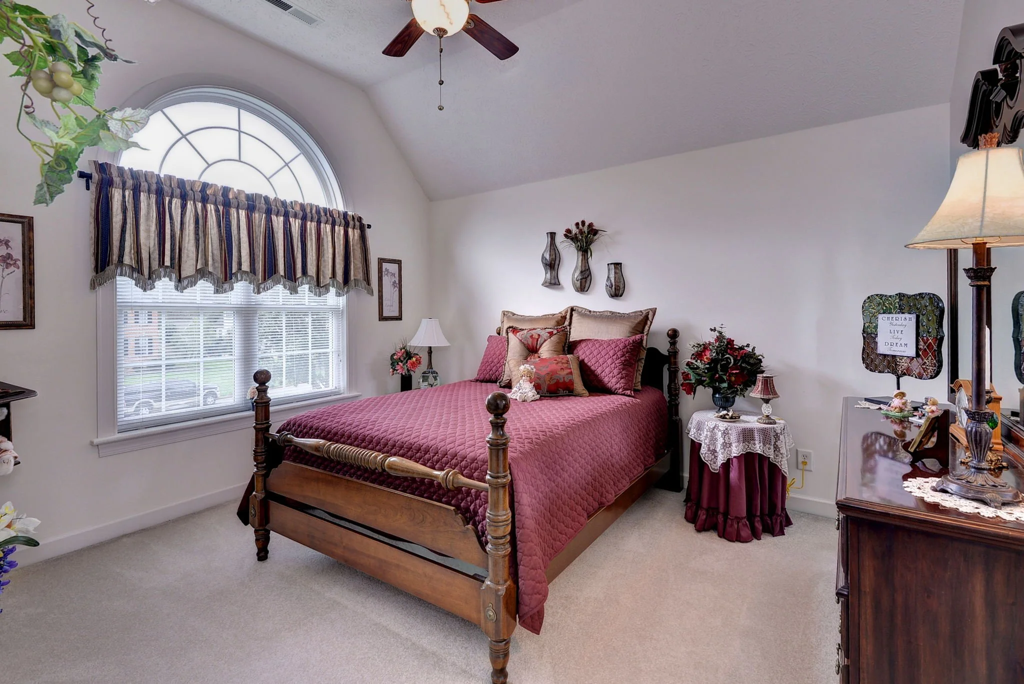 A bedroom with a large wooden bed featuring a pink quilt and multiple pillows. There is a window with striped curtains, a ceiling fan, and a nightstand with a flower arrangement and lamp. The room is decorated with framed artwork and wall-mounted vas