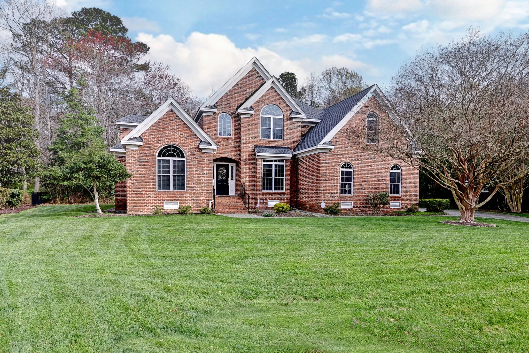 A large brick house with multiple gabled roofs, tall arched and rectangular windows, a black front door, and a well-maintained green lawn with trees surrounding the house.