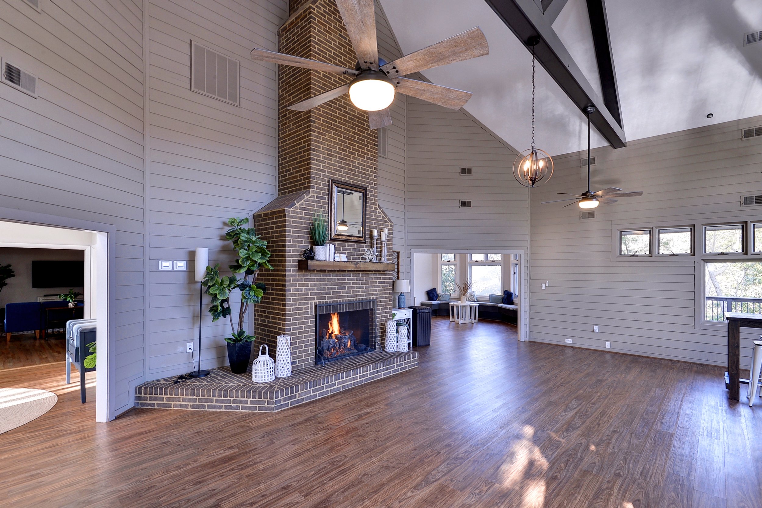 Spacious living room with a high vaulted ceiling, white wood paneled walls, a brick fireplace with a fire burning, ceiling fans, and large windows letting in natural light. Decor includes potted plants, candles, and wall art.