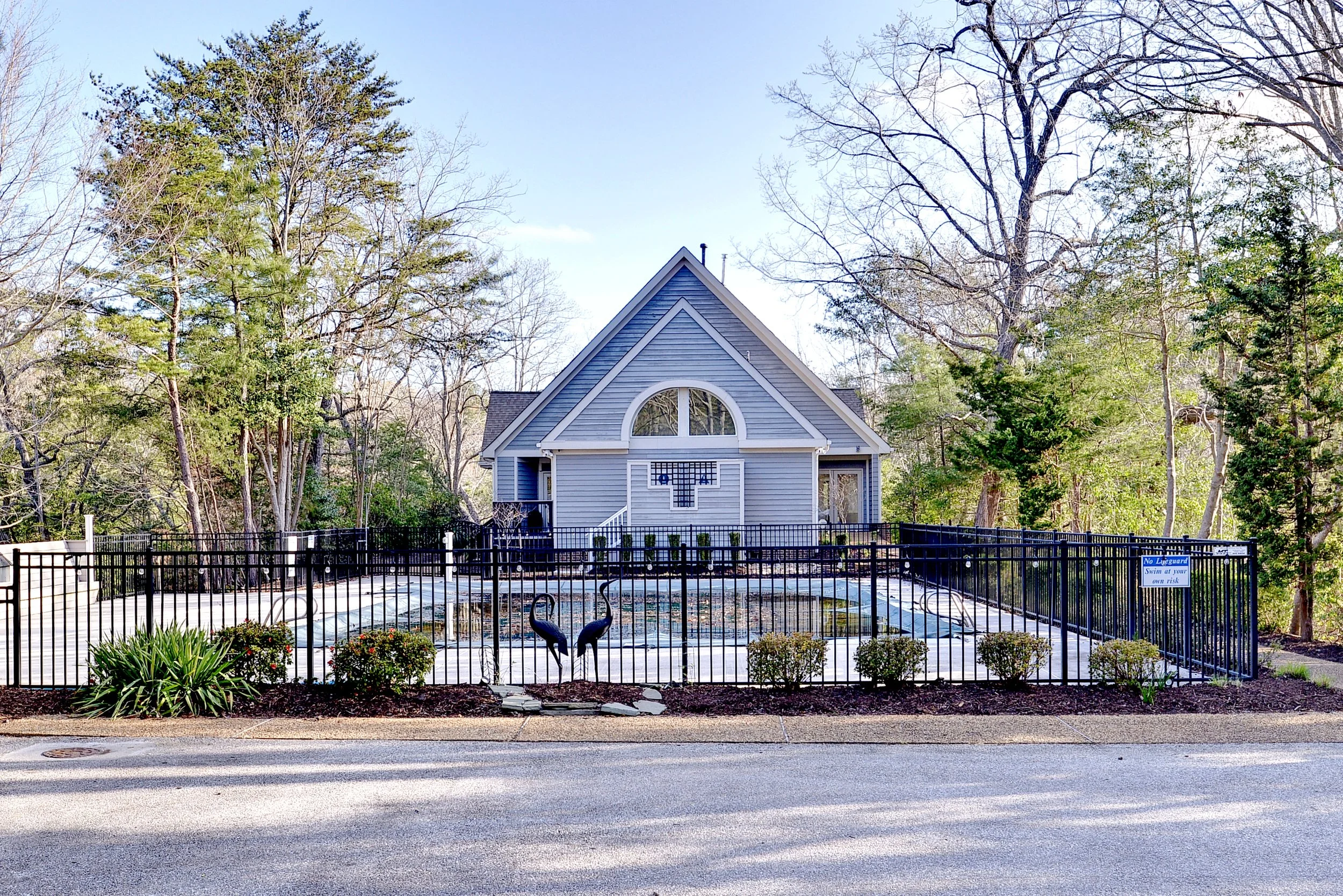 A house painted in light gray with a steep front-facing gable, surrounded by trees and a black metal fence enclosing a pool with a small garden in front.