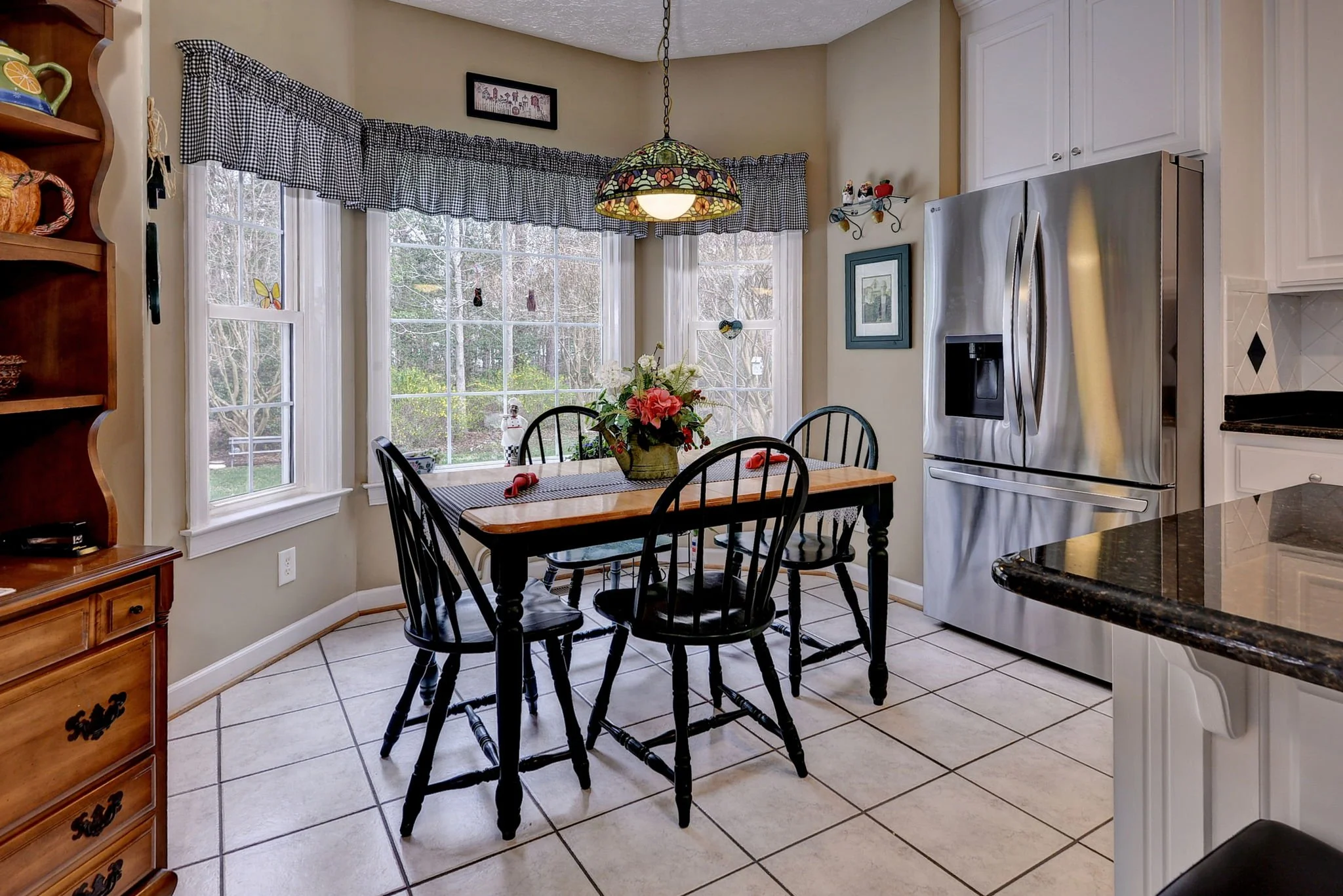A cozy kitchen dining area with a wooden table, black chairs, a ceiling stained glass lamp, a flower centerpiece, large windows with black and white checkered curtains, and stained glass decorations on the window. There's a stainless steel refrigerat