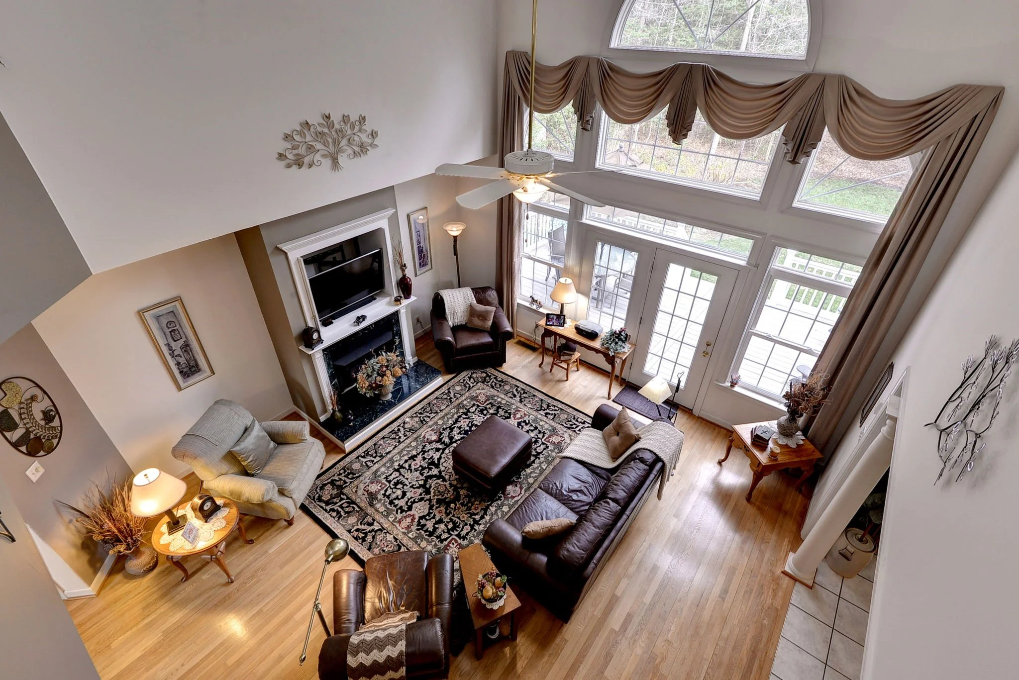 Living room with large windows, beige curtains, and hardwood floors. Contains a brown leather sofa, a beige armchair, an ottoman, a fireplace with a TV above, tables with lamps, and various decorative items.