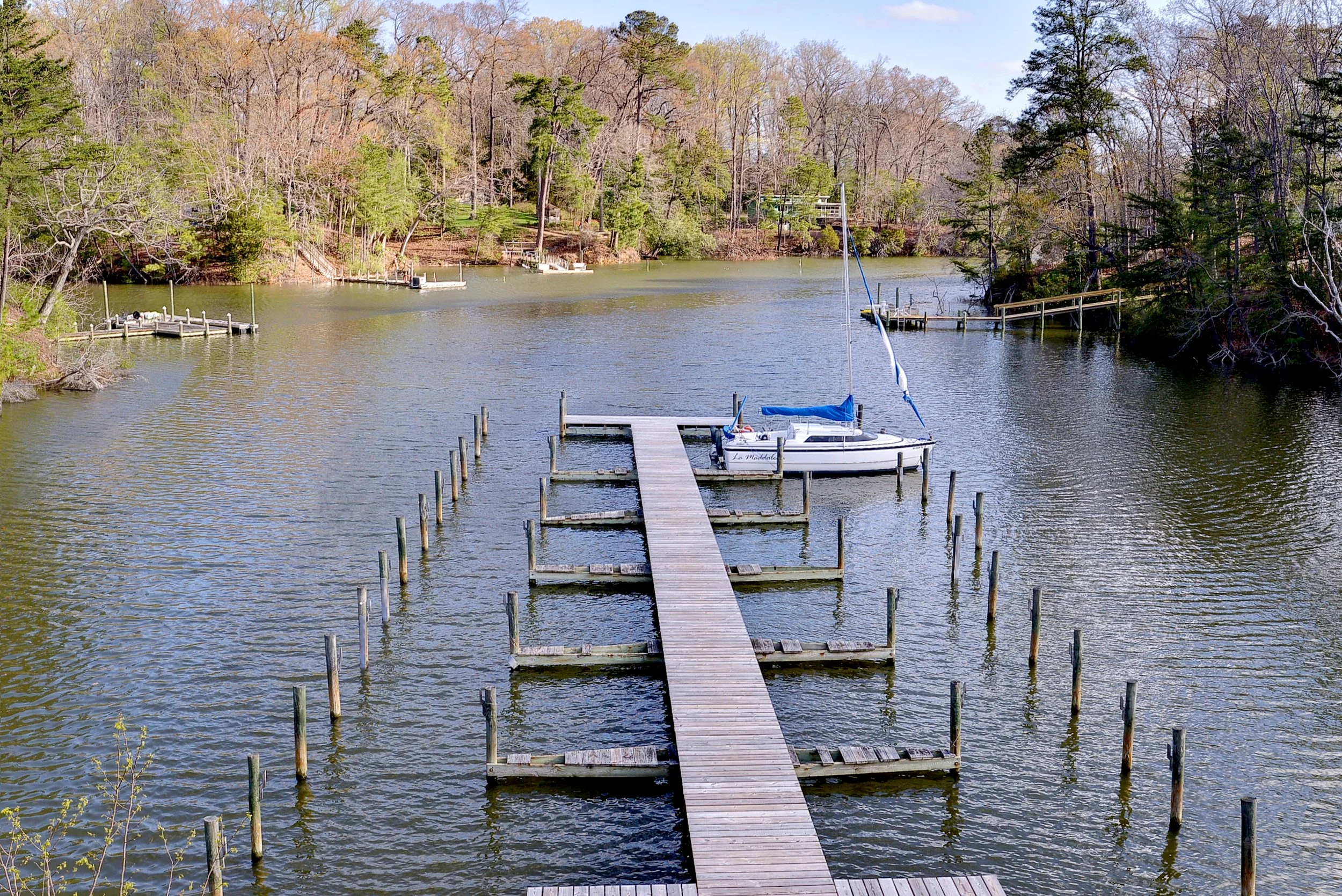 A small marina with a wooden dock extending into a calm lake, where a sailboat is moored. The background features trees with some greenery and some without leaves, indicating early spring or late fall.