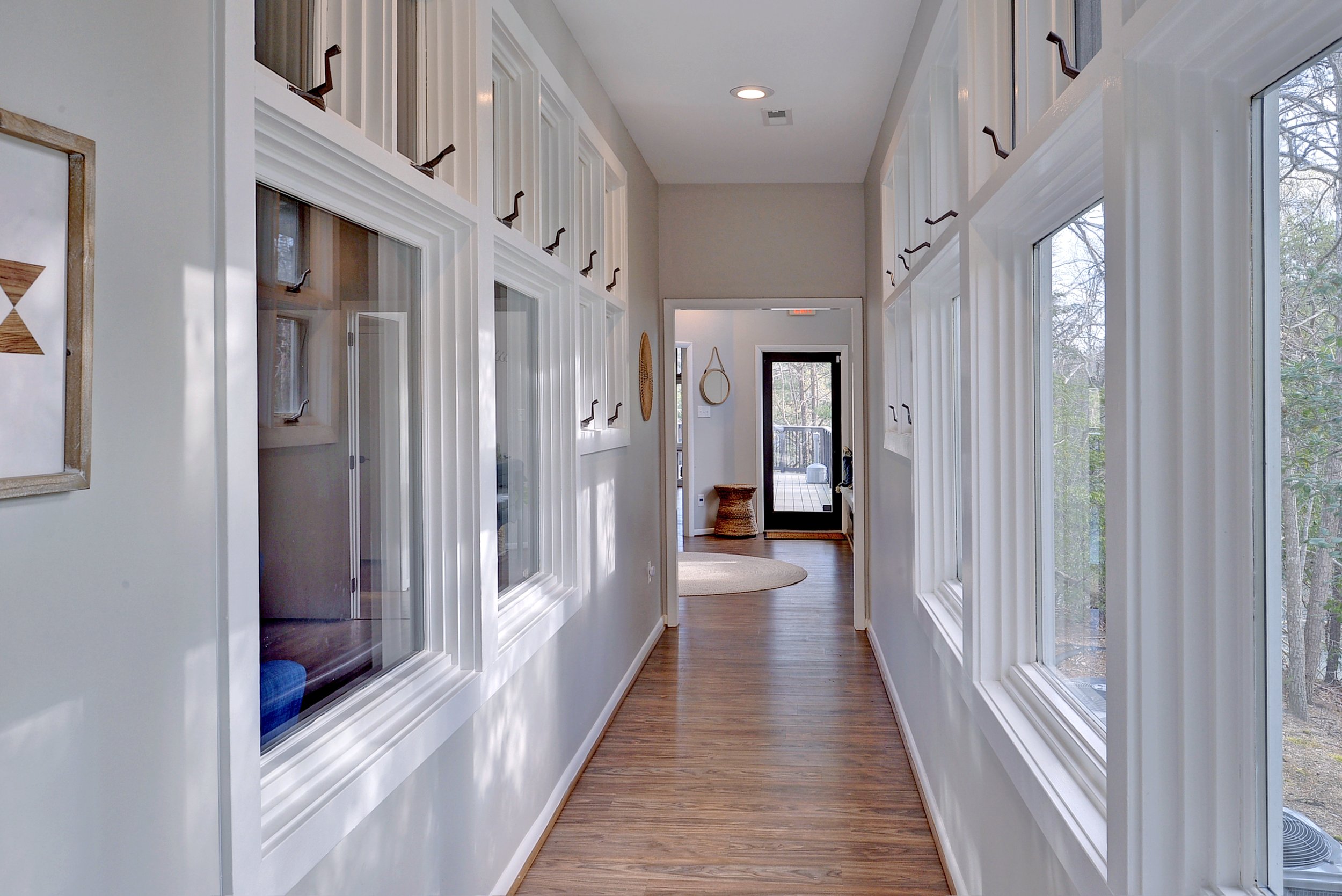 Indoor hallway with white walls, hardwood floors, multiple windows on both sides, and decorative hooks along the top of the walls. Leading towards a door with a glass panel, revealing an outdoor deck with trees.