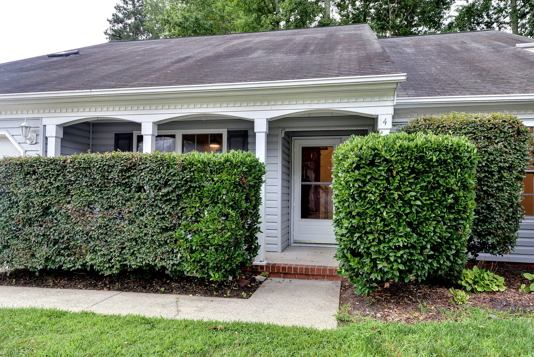 Front view of a house with gray siding and a dark shingle roof, partially concealed by green bushes and trees.