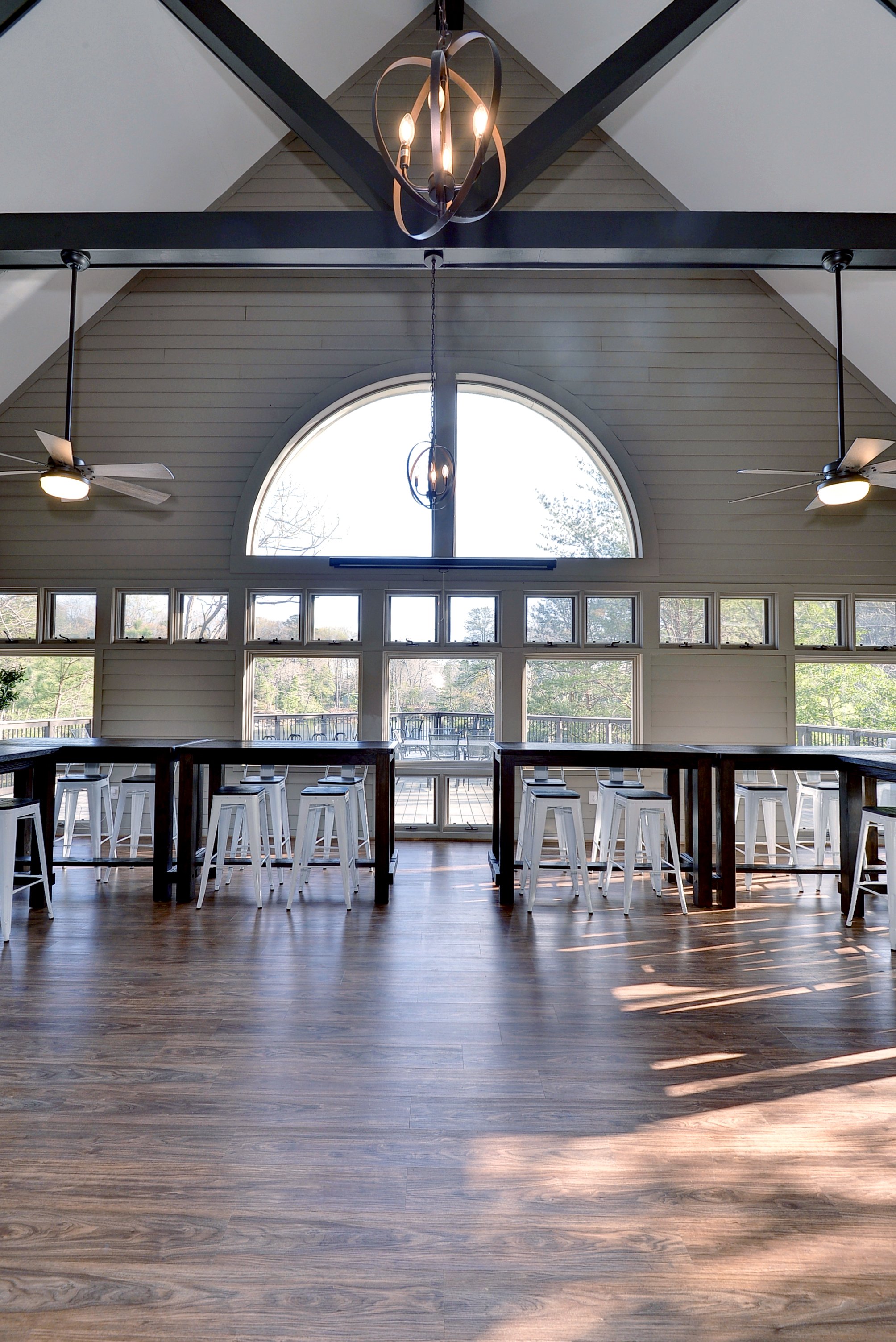 Interior of a bright room with large arched window, ceiling fans, and a chandelier, featuring two high tables with white stools, in a room with wooden floors and a view of trees outside.