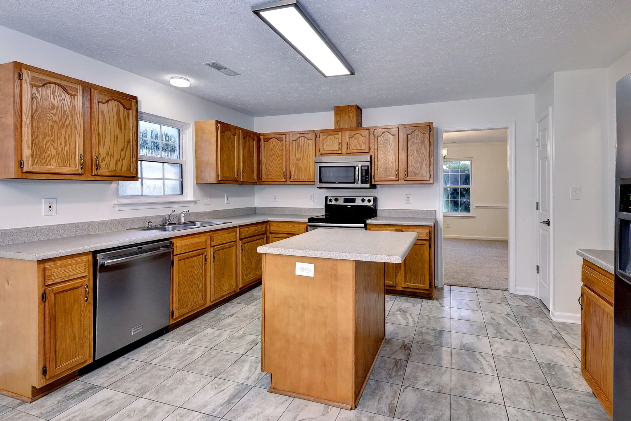 A kitchen with wooden cabinets, a countertop, a window, a stove, a microwave, and tiled flooring.