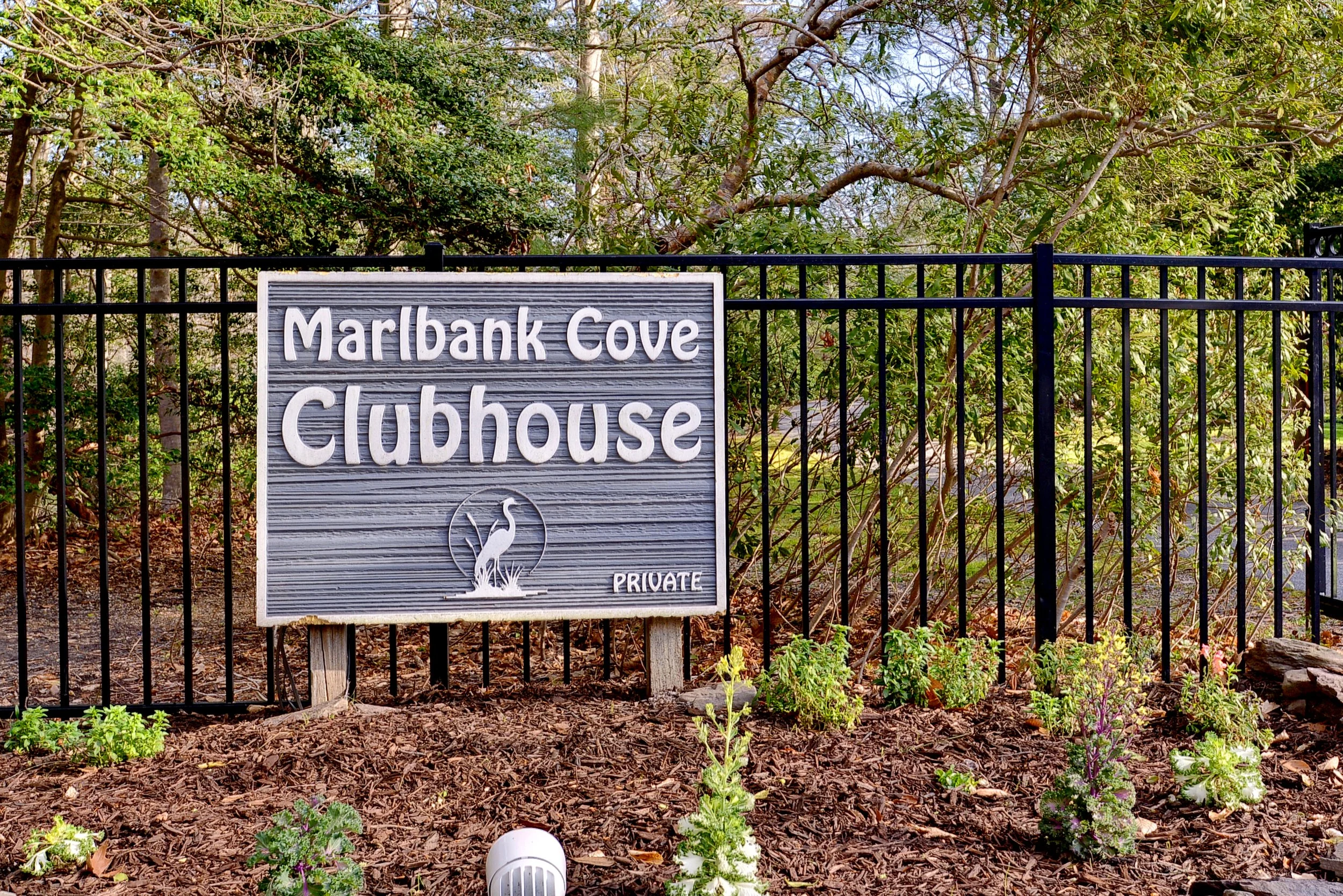 Sign for Marlbark Cove Clubhouse with a bird illustration, set against a greenery background and a black metal fence