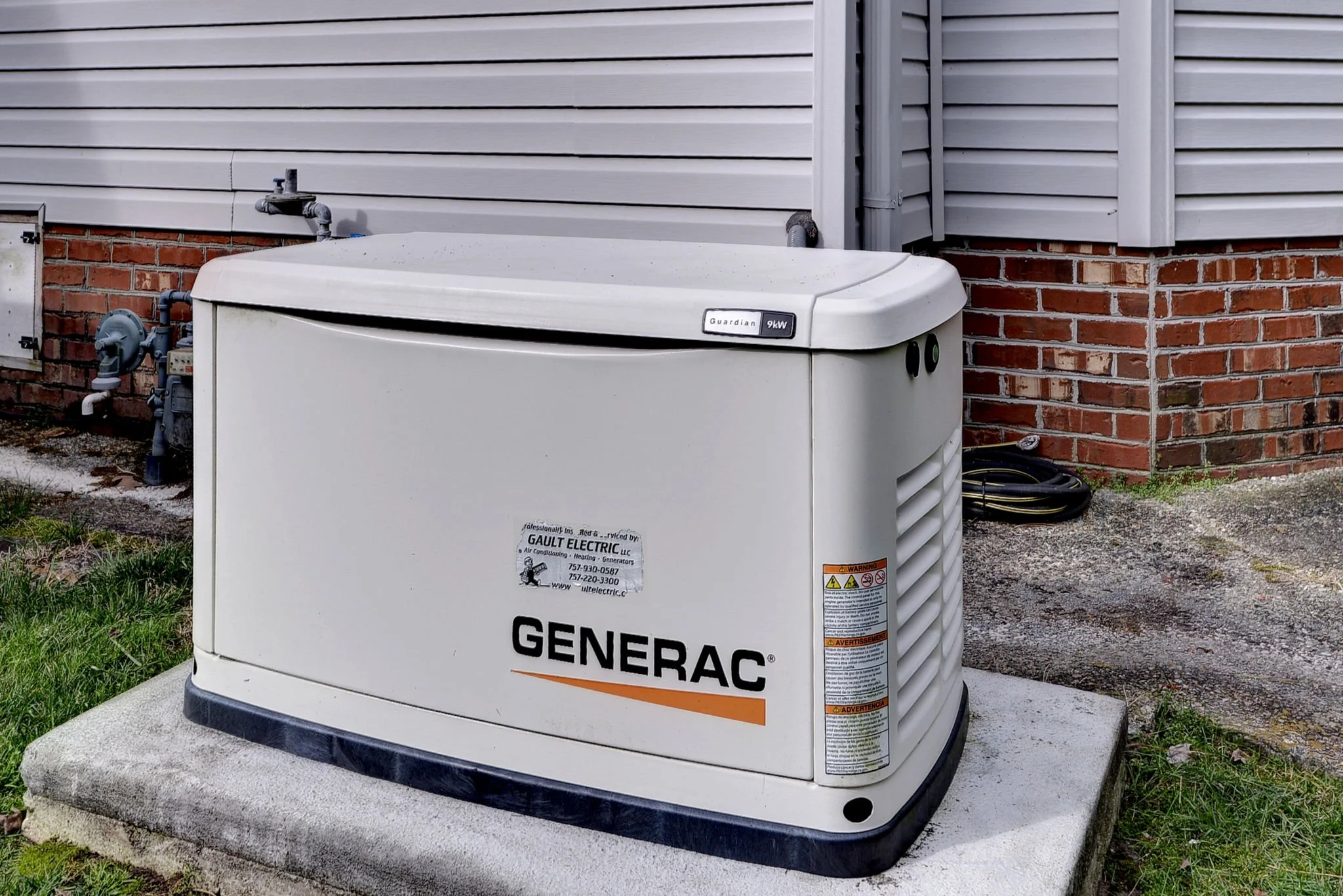 A white portable generator with the brand name 'GENERAC' on it, placed on a concrete pad outside a house with gray siding and red brick foundation.