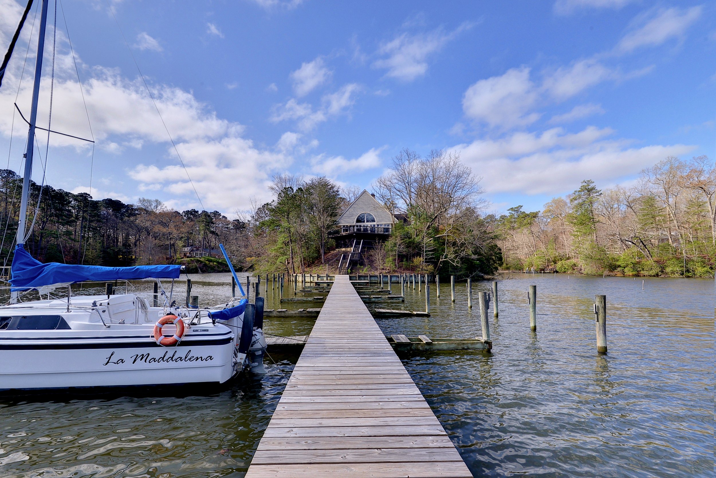 A wooden dock extending over a body of water towards a house on a hill, with a sailboat named La Maddalena moored on the left and trees on both sides under a partly cloudy sky.