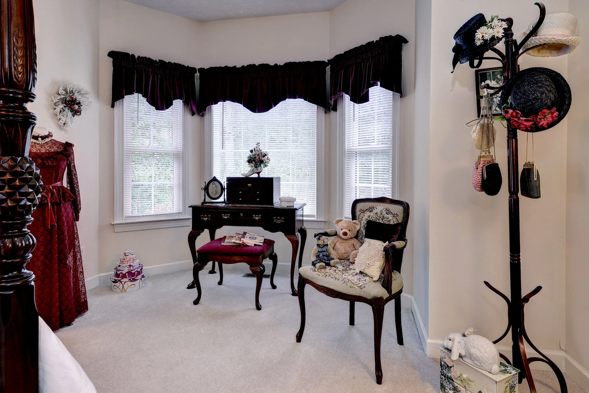 A cozy corner of a room with a bay window, decorated with vintage furniture, including an ornate vase of flowers on a small table, a velvet upholstered chair with stuffed animals, a black dresser with a round mirror, and a tall coat rack with hats an