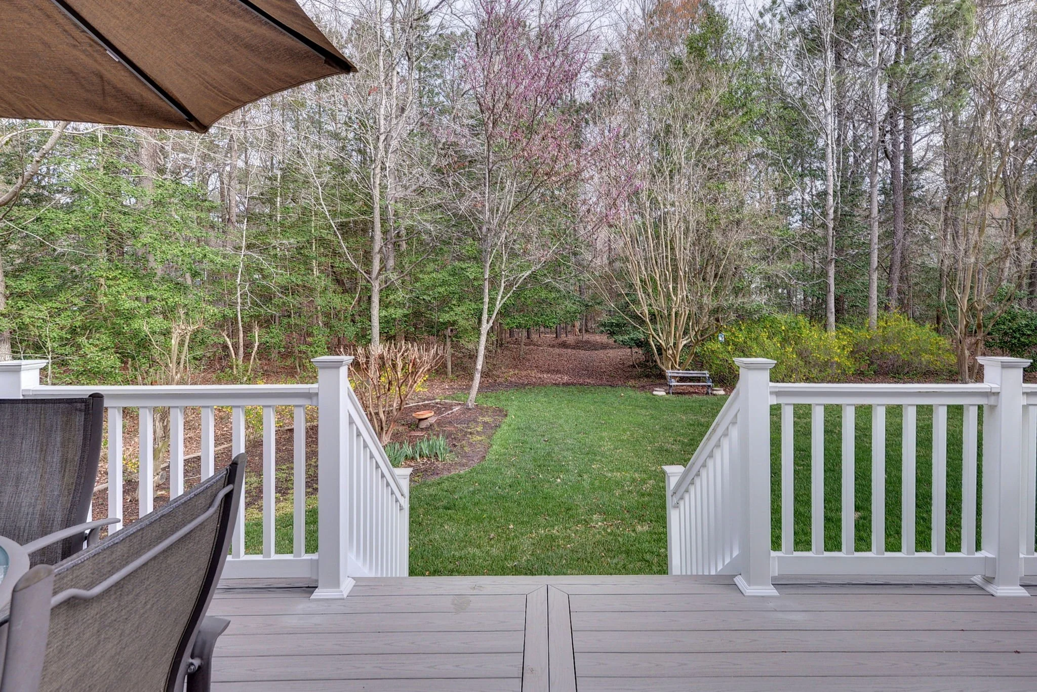 View of a backyard with a wooden deck, white railing, green grass, and trees with bare branches and some green foliage, seen from a porch with outdoor furniture and a large umbrella.
