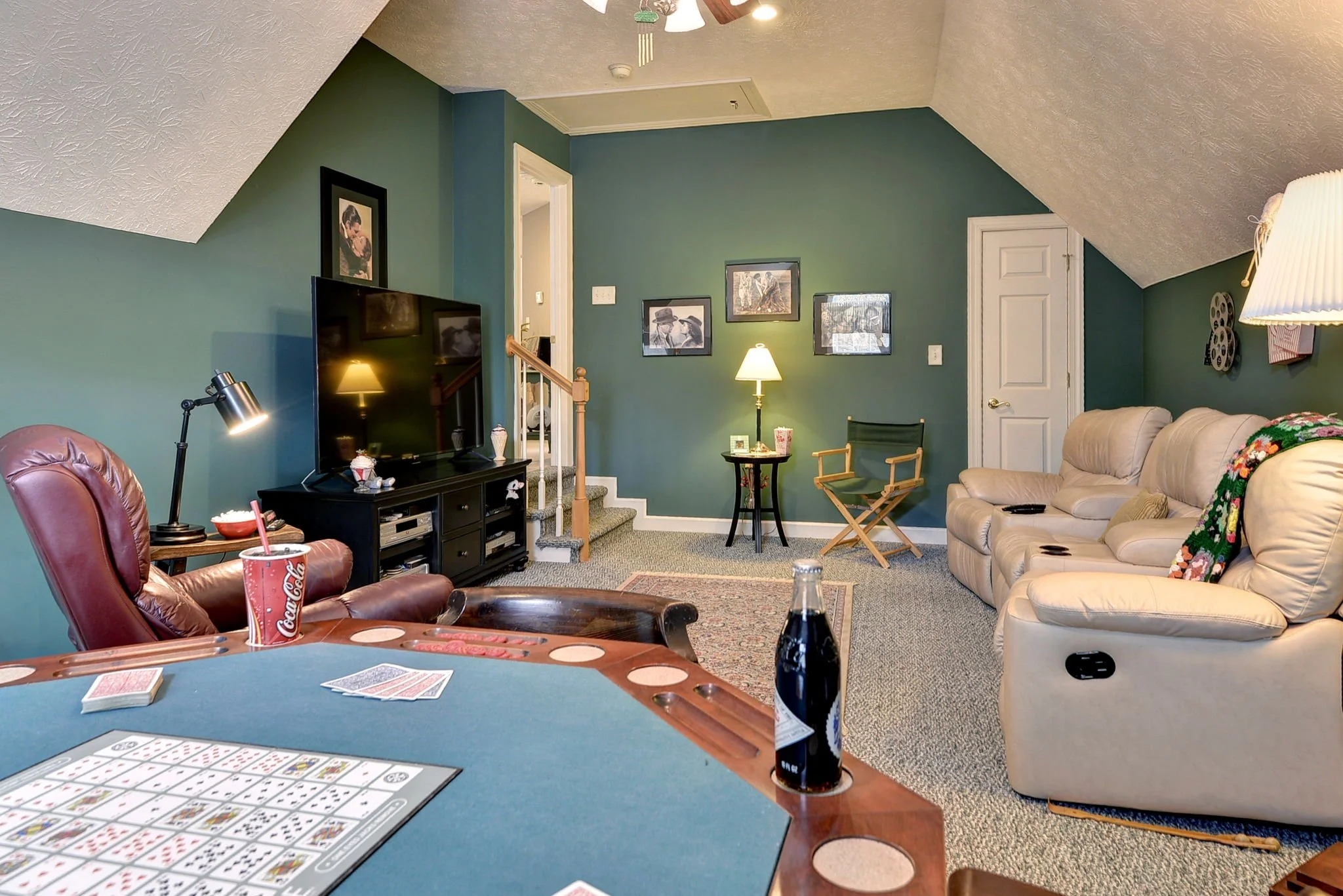 A living room with a green accent wall, beige sofa, and a TV on a black stand. A game of bingo is in progress on a round table in the foreground, with drink cups and a Coke bottle. There are framed pictures on the wall, a standing lamp, and a directo