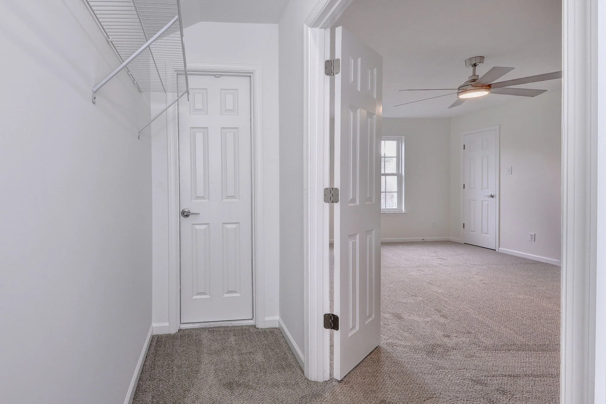 Interior view of a modern home hallway with white walls, carpeted floors, and multiple doors leading to different rooms. A ceiling fan is visible in one of the rooms.