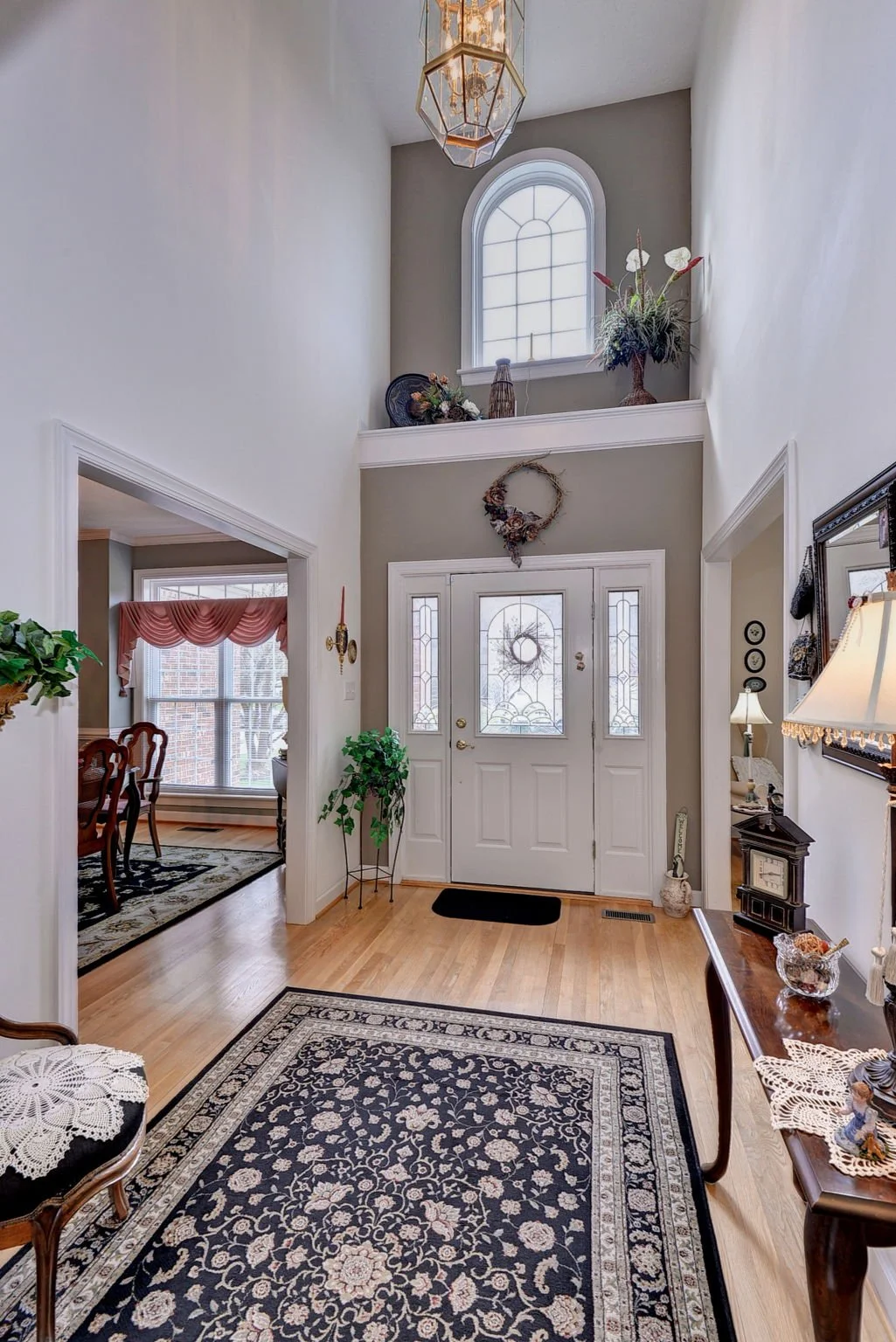 View of a well-lit foyer with a front door, decorative glass panels, and an arched window above. The space features a patterned rug, plants, framed art, and vintage furniture, with a staircase and upper gallery decorated with plants and vases.