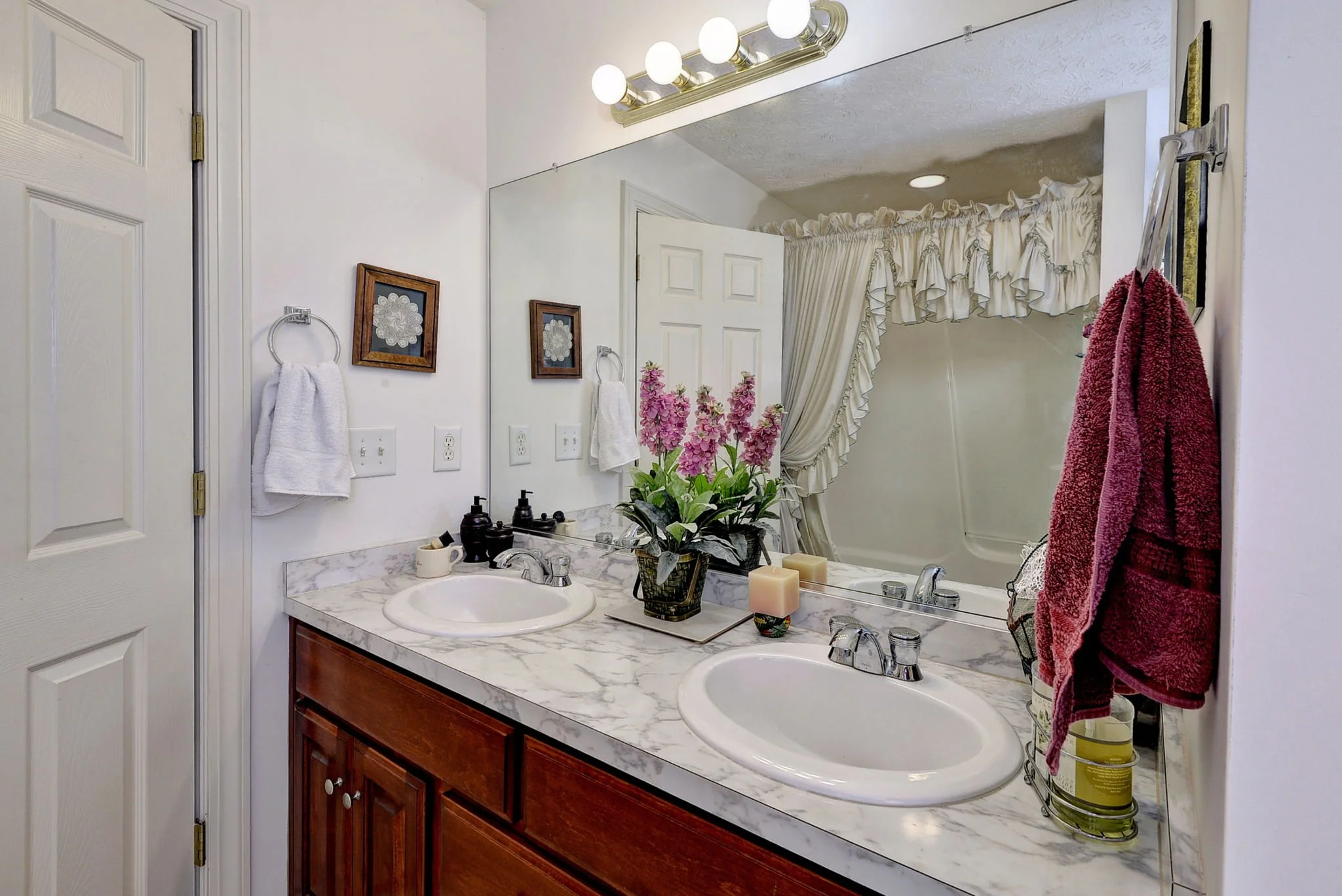 A bathroom with a marble countertop, double sinks, a large mirror, and a cabinet below. There are plants, candles, and soap dispensers on the counter. Towels are hanging on rings on the wall, and there are framed pictures and a robe or towel hanging 