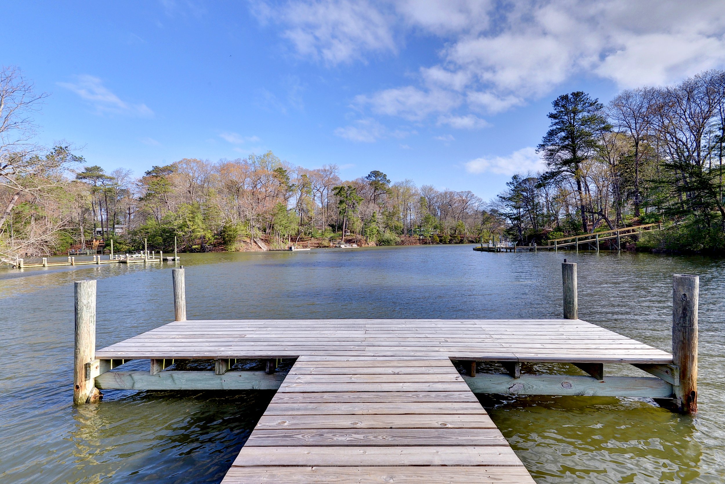 A wooden dock extending into a calm lake with trees in the background, under a blue sky with some clouds.