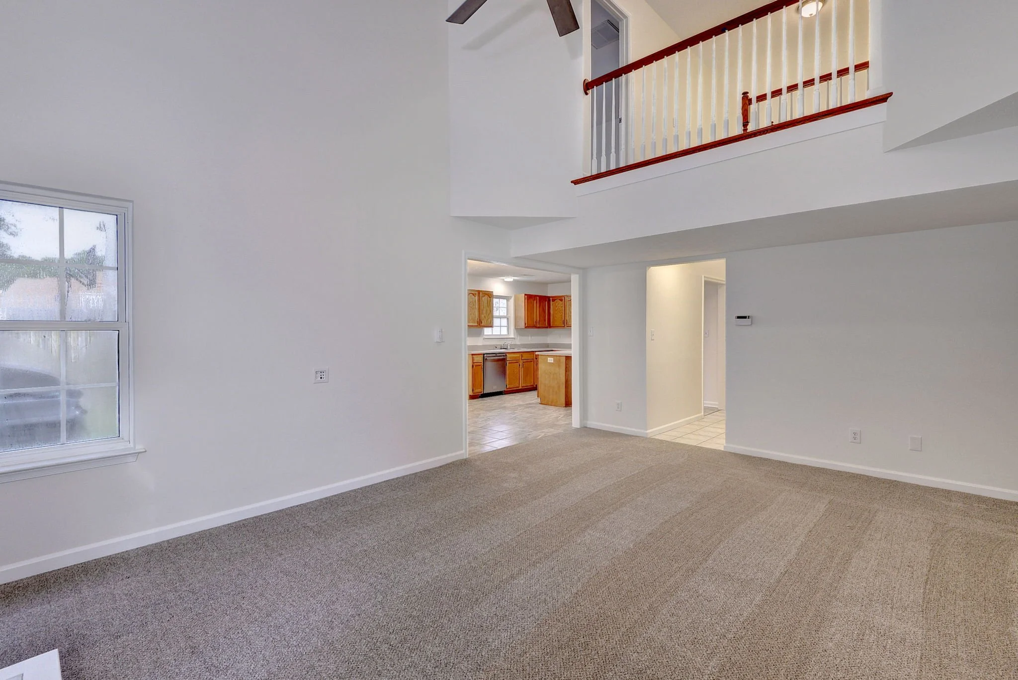 Living room with beige carpet, white walls, a window, an open kitchen with wooden cabinets, and a staircase with a wooden railing leading upstairs.