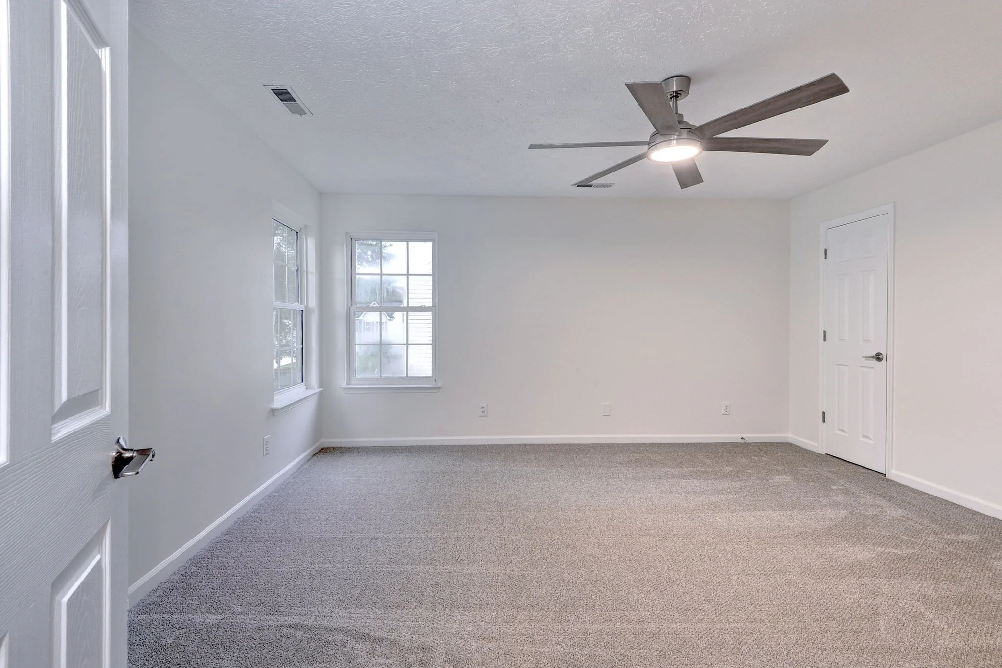 Empty room with white walls, carpeted floor, two large windows, a ceiling fan, a door, and multiple electrical outlets.