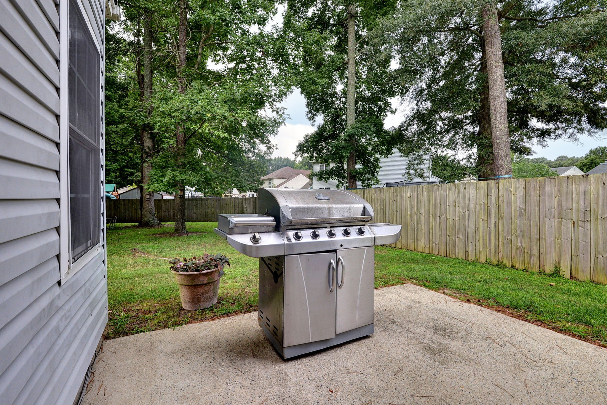 Backyard patio area with a stainless steel outdoor grill, potted plants, and a wooden fence, surrounded by green trees and neighboring houses.