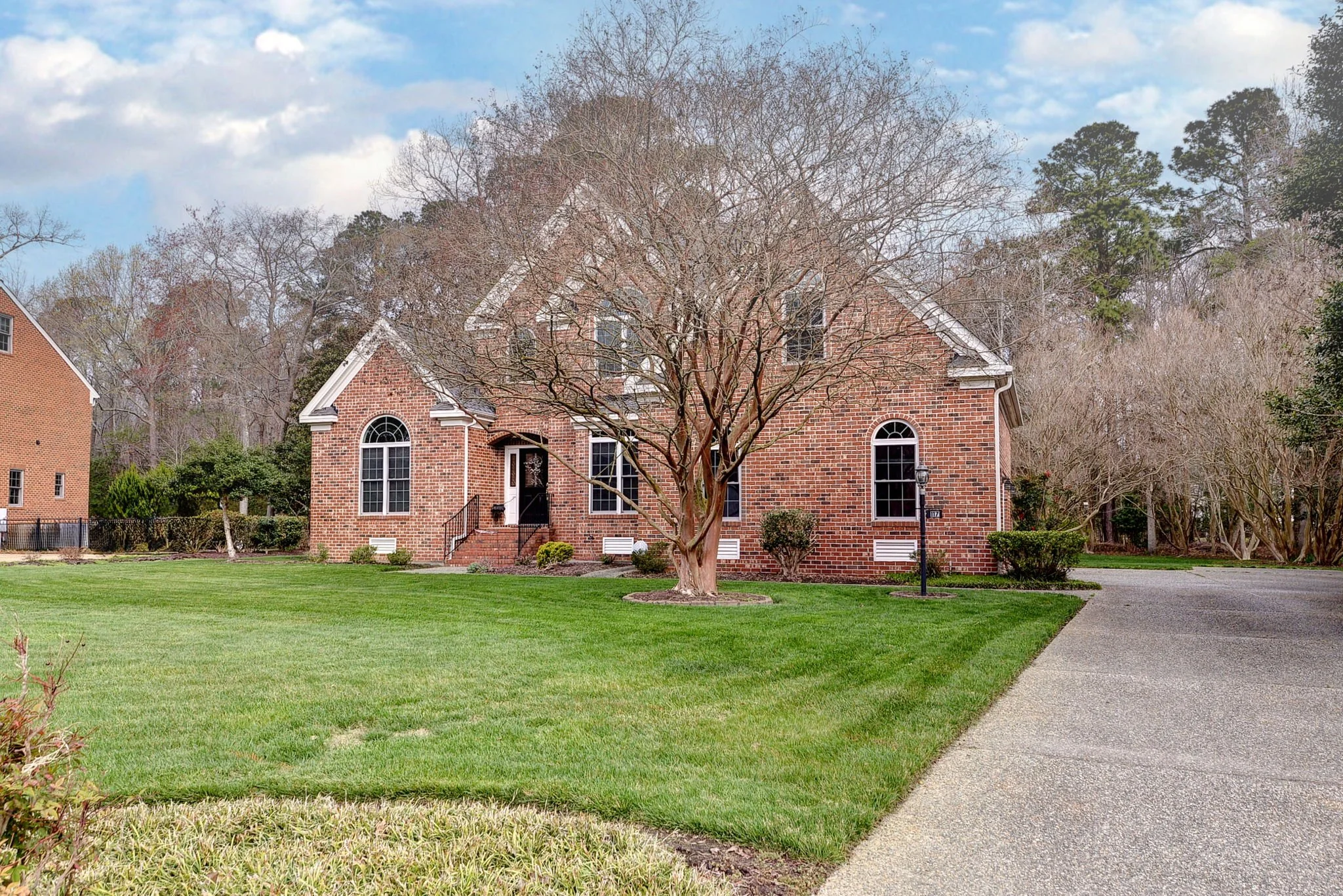 front view of a brick house with large arched windows, a tree with no leaves in the front yard, green lawn, and a curved driveway under a partly cloudy sky