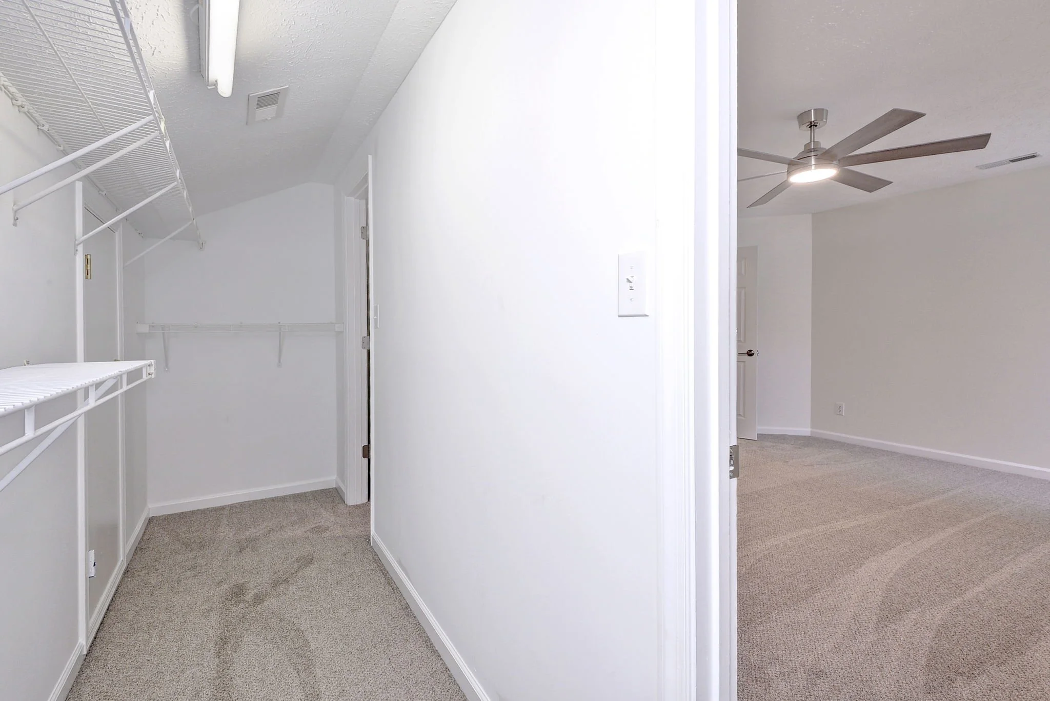 A small room with white wire shelving on the left wall, beige carpeted floor, and a white door at the back. The adjacent room has a ceiling fan and beige carpet.