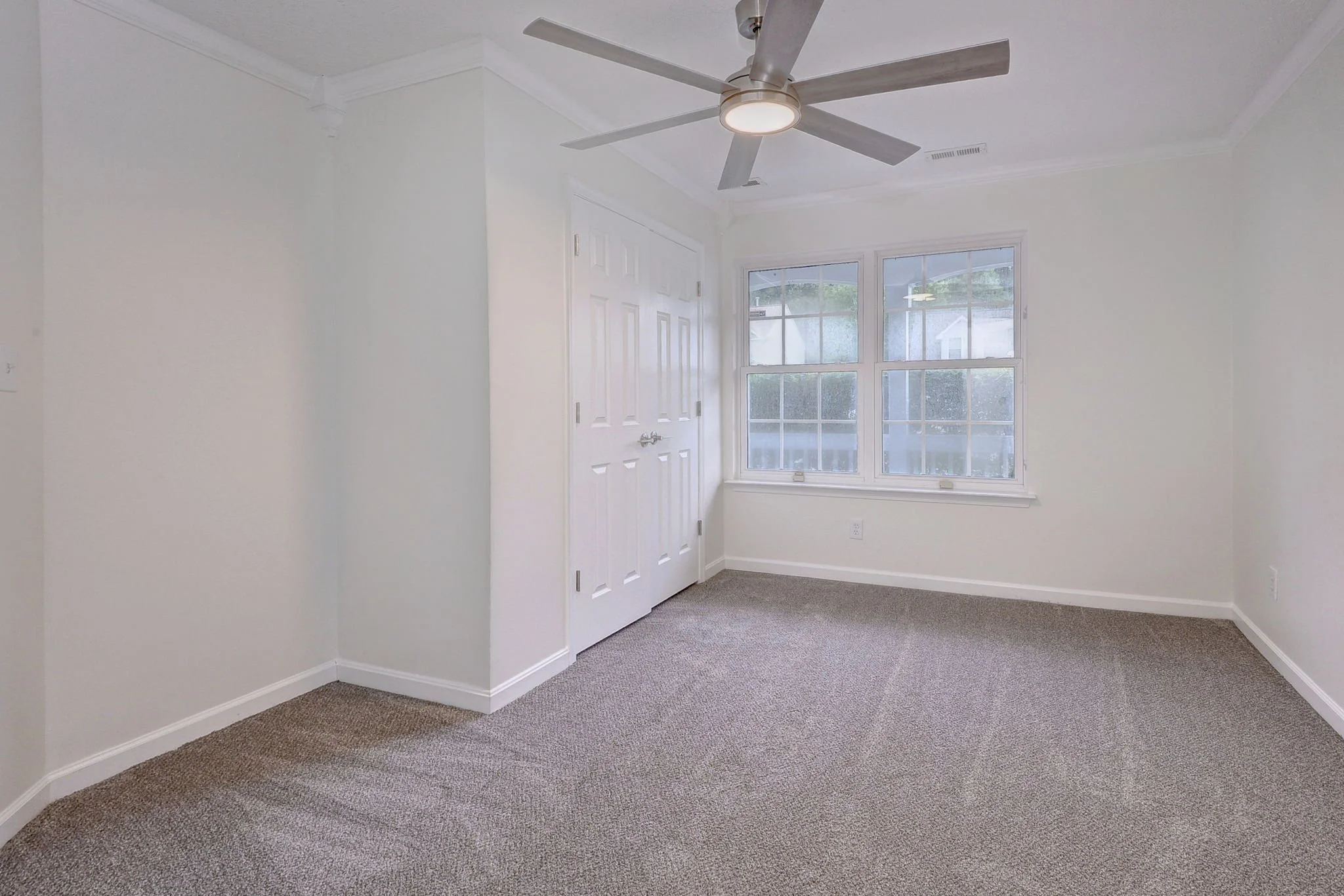 A bright, empty room with beige carpet, white walls, a ceiling fan, a window, and a closet with white double doors.