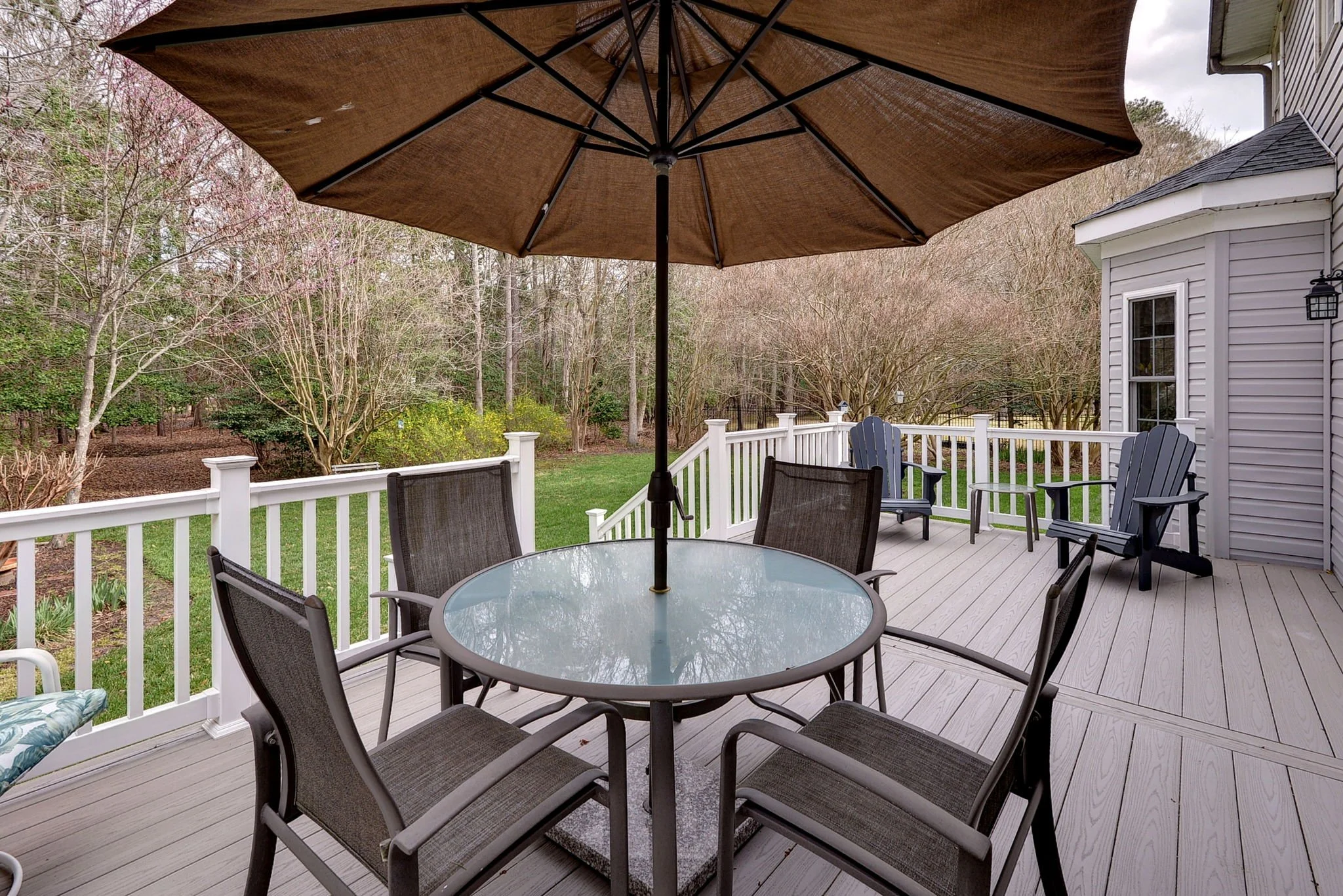 Outdoor deck with a round glass table, six chairs, large umbrella, Adirondack chairs, and trees in the background.