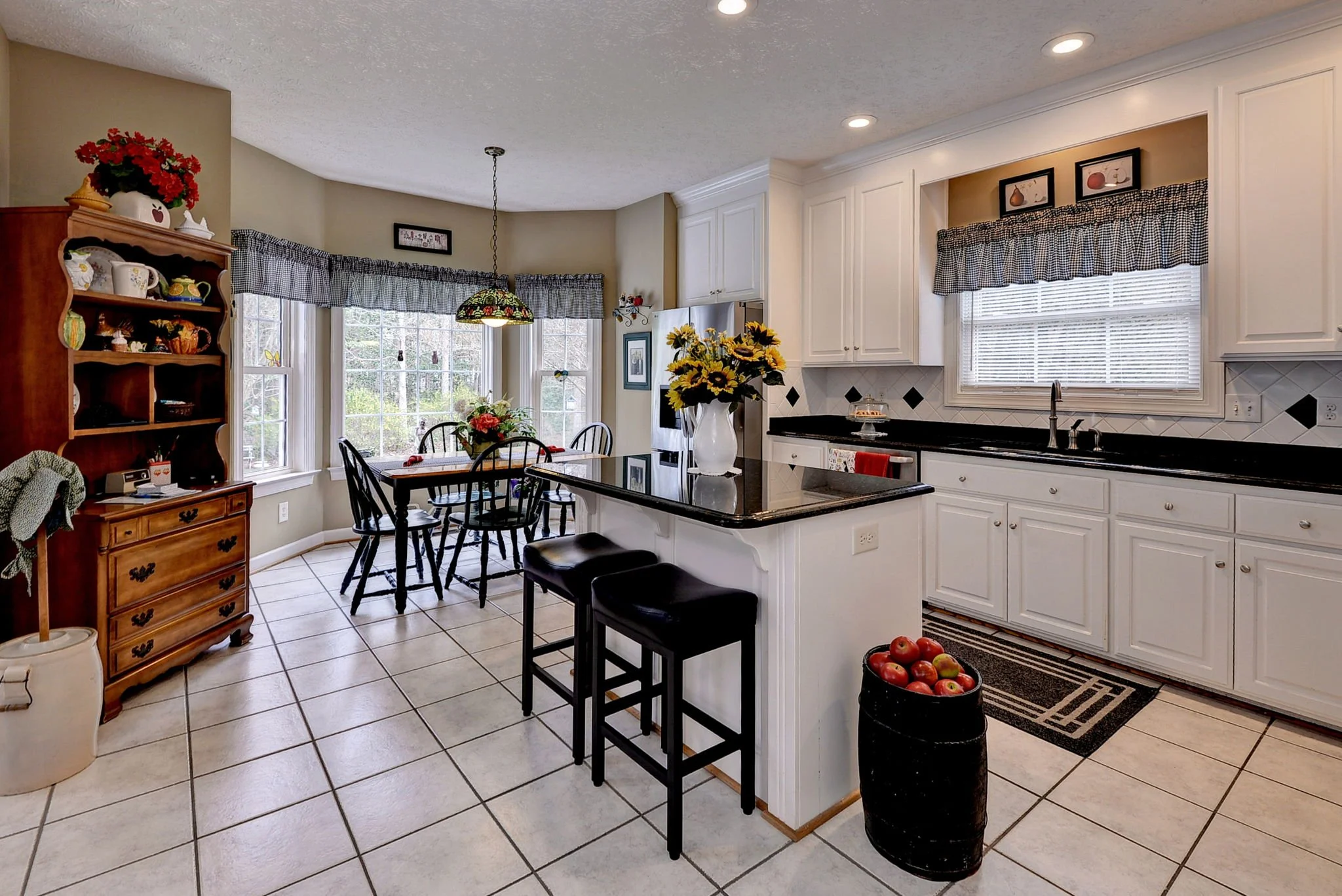 A kitchen with white cabinets, black countertops, and a black and white tiled floor. There is a dining area with a wooden table, four black chairs, and a stained glass pendant light. The room has large windows with checkered valances, and a wooden hu