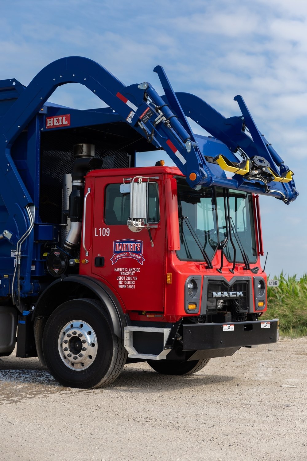 A red and blue garbage truck with a hydraulic lift arm, parked on a gravel surface under a partly cloudy sky.