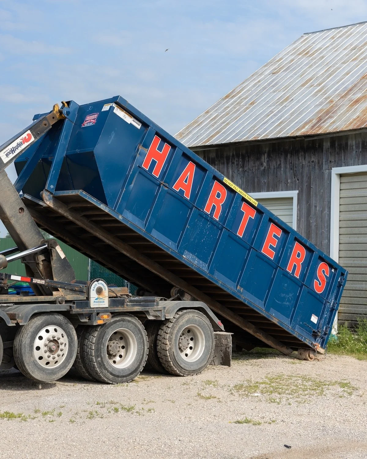 A blue dump trailer with the word "HARTERS" painted in red on the side, raised at an angle, parked on a gravel surface near a wooden building with a metal roof.