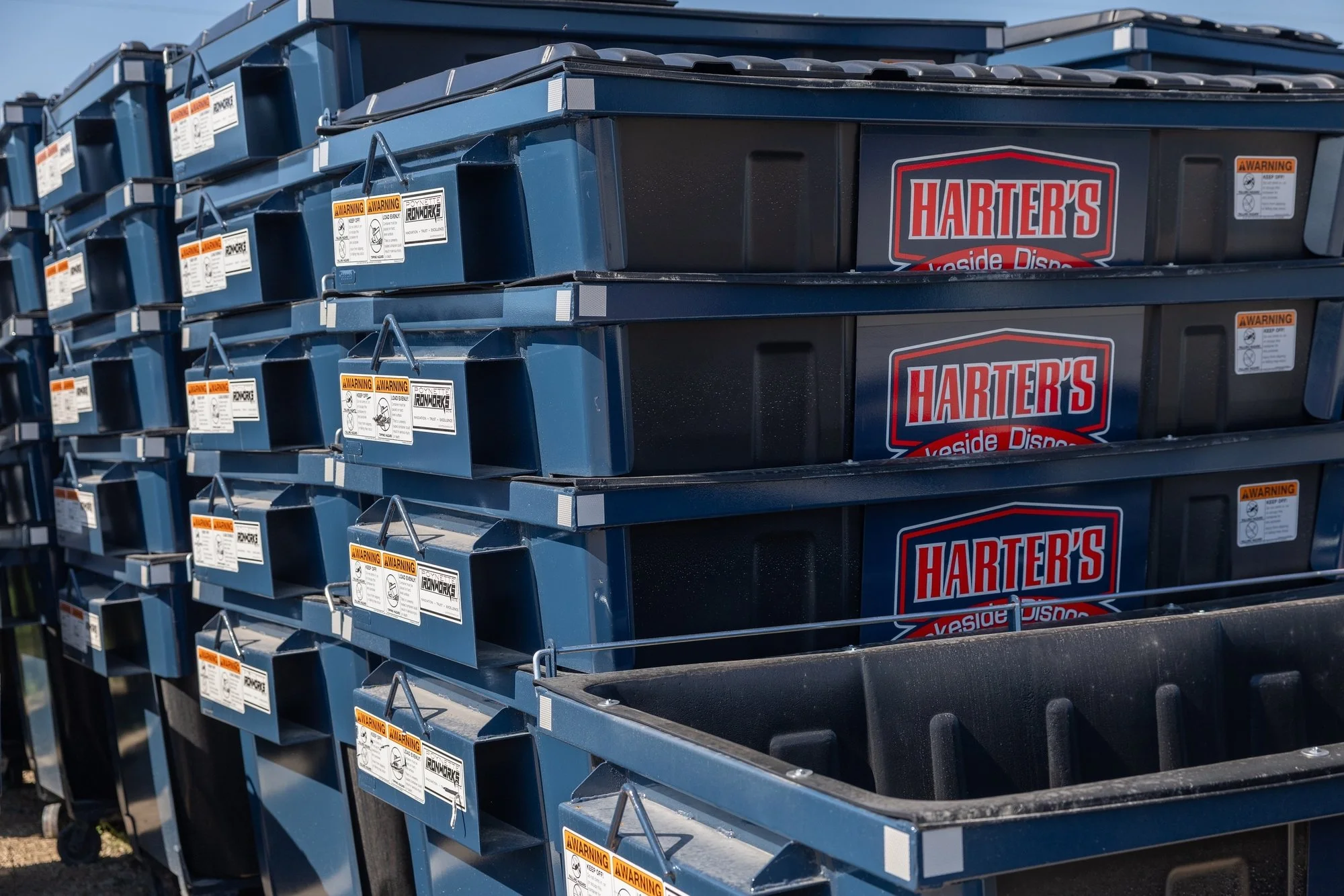 Stacked blue plastic storage bins with the label 'Harter's' visible on some of them, outdoors under bright sunlight.