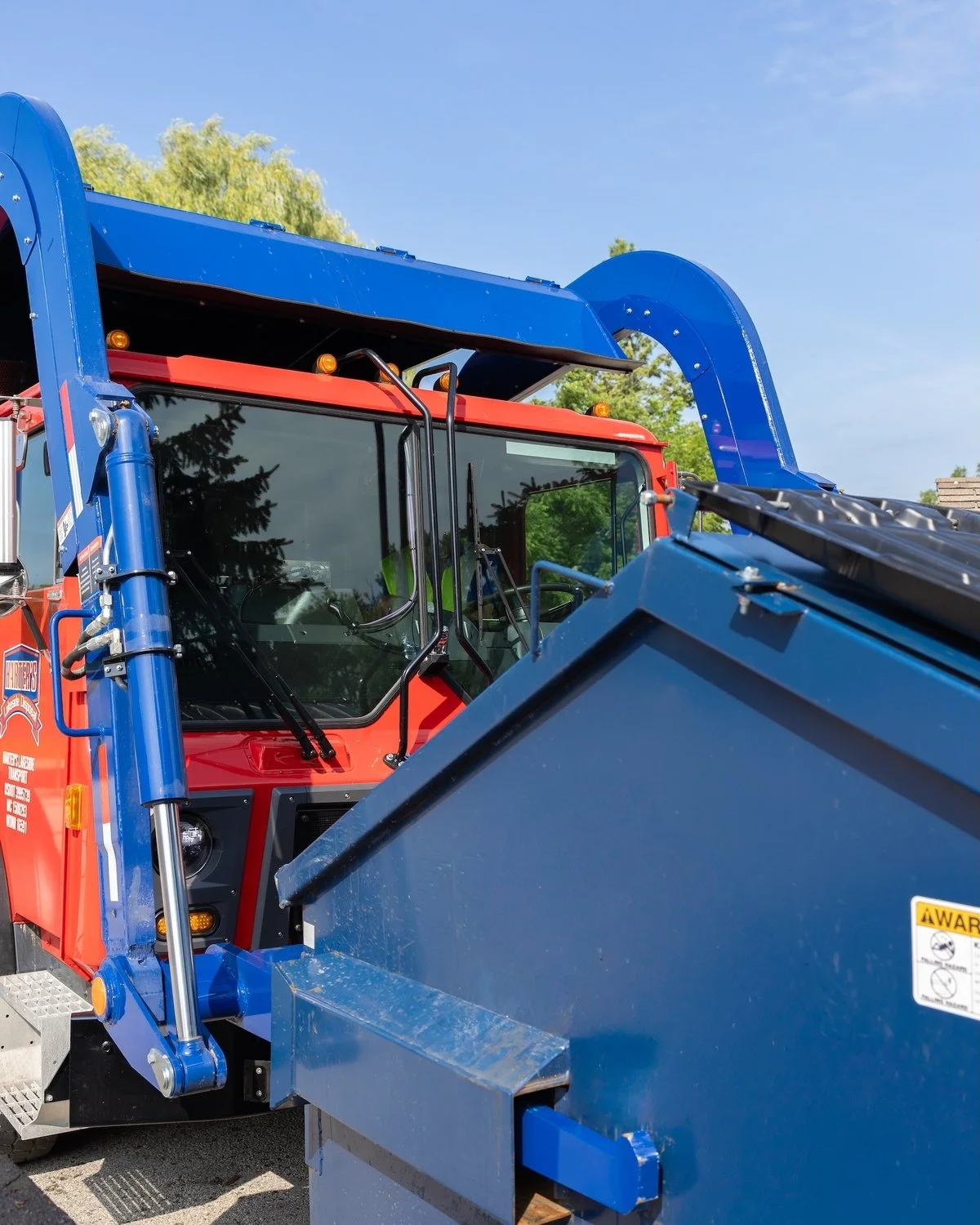 Close-up of the front part of a fire truck with a blue fire hose reel and a blue debris container, red cab, and clear sky.