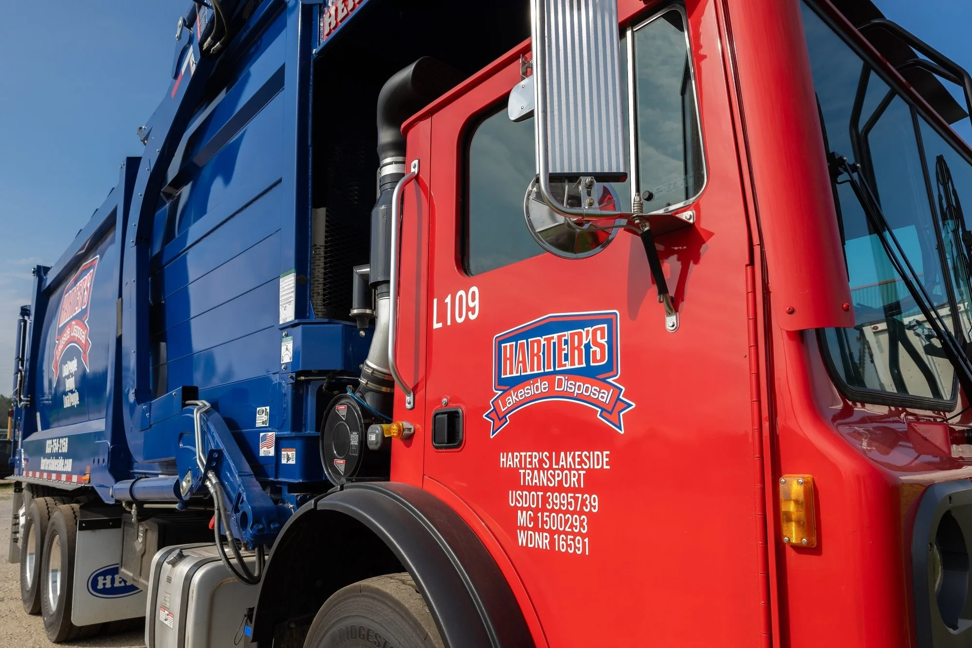 A colorfully painted garbage truck with the logo for Harter's Lakeside Disposal on the side, featuring a red cab and blue body, parked under a blue sky.
