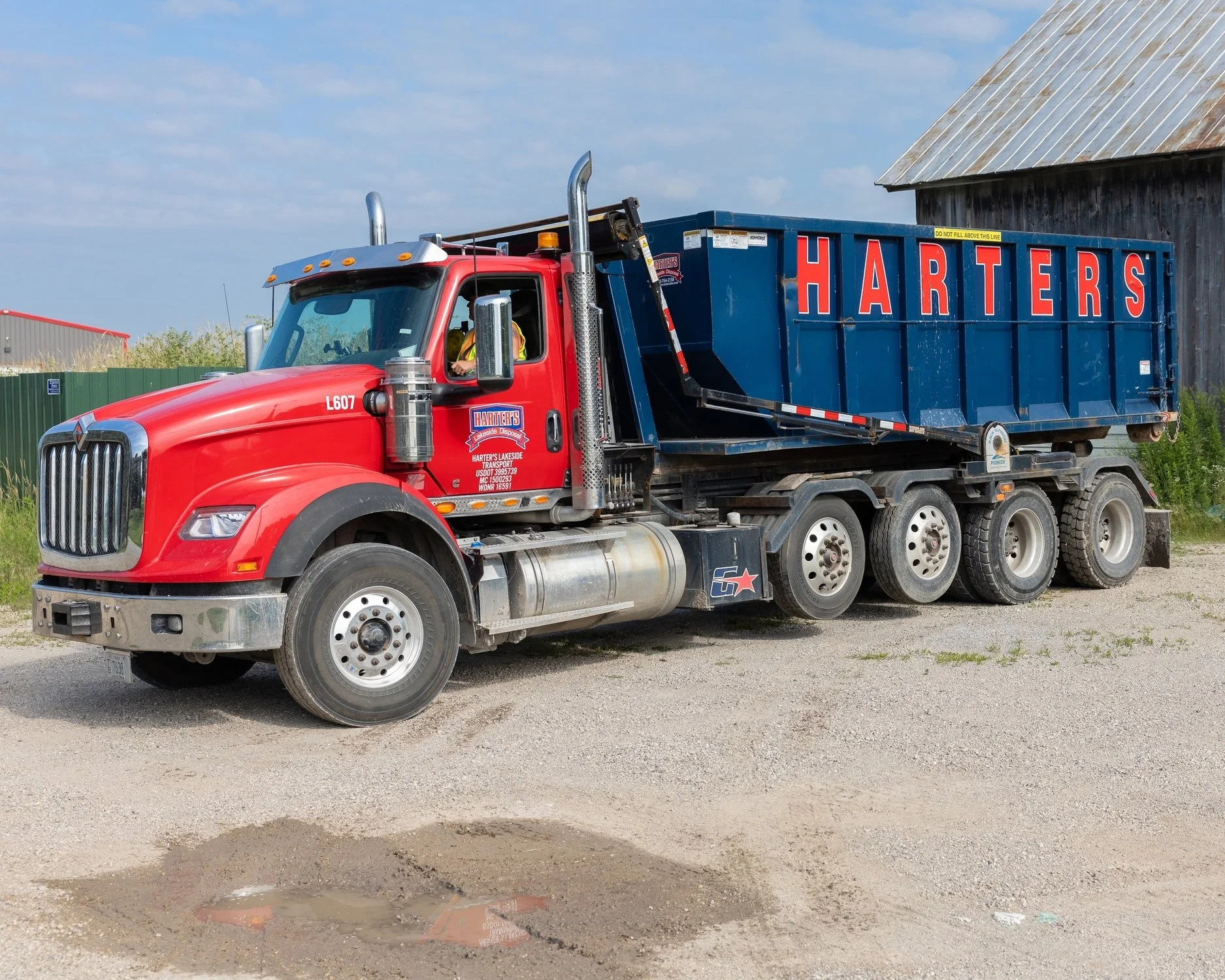 Red dump truck with blue container and red lettering that says 'HARTERS', parked on gravel, with a weathered barn in the background.