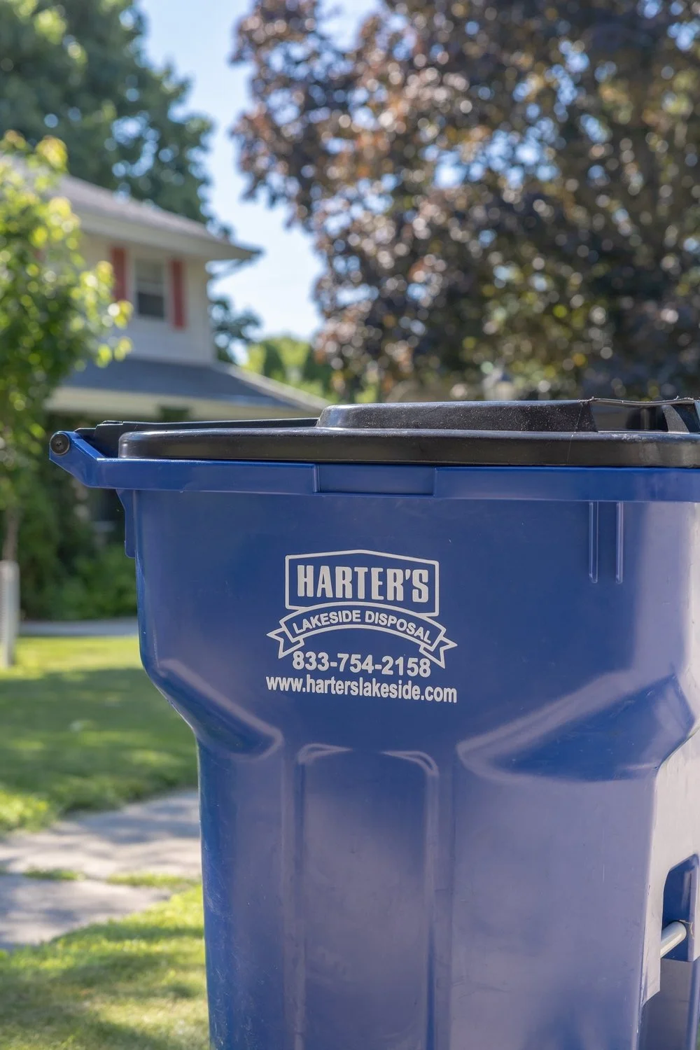 A blue waste bin with Harter's Lakeside Disposal logo, phone number, and website, outdoors in a residential neighborhood with trees and a house in the background.