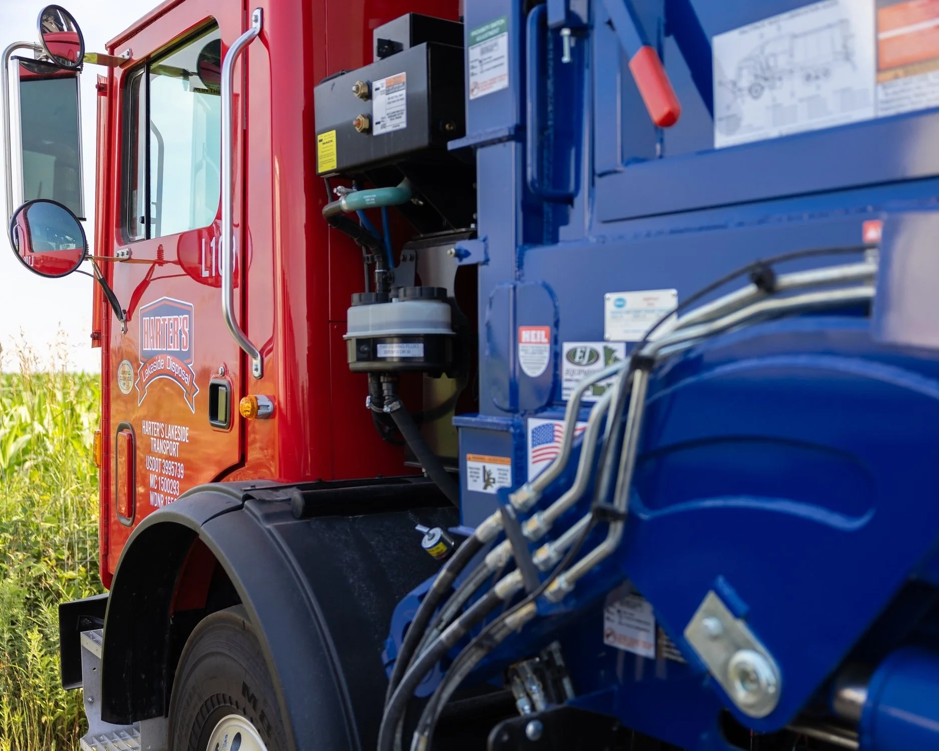 Close-up of a fire truck's side with visible hoses, control panel, and mirrors, parked in a rural area with greenery in the background.