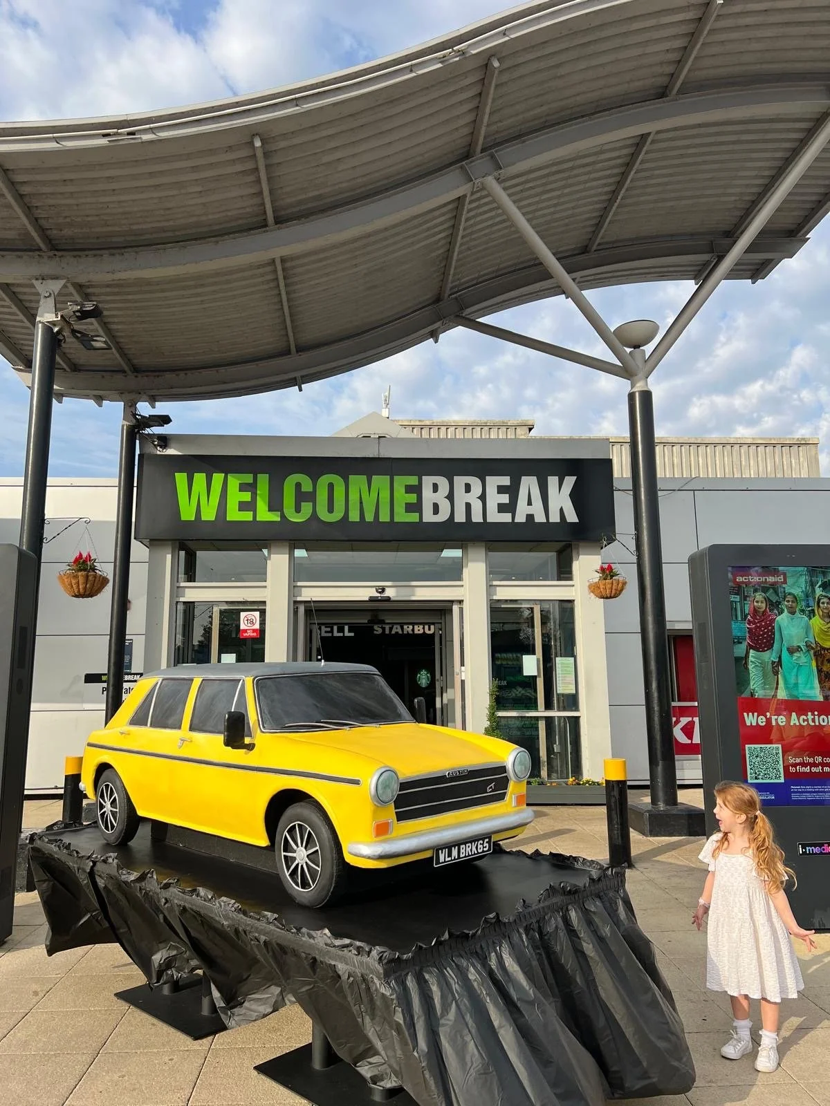 A young girl in white dress admiring a giant yellow car cake in front of Welcome Break Motorway Service Station