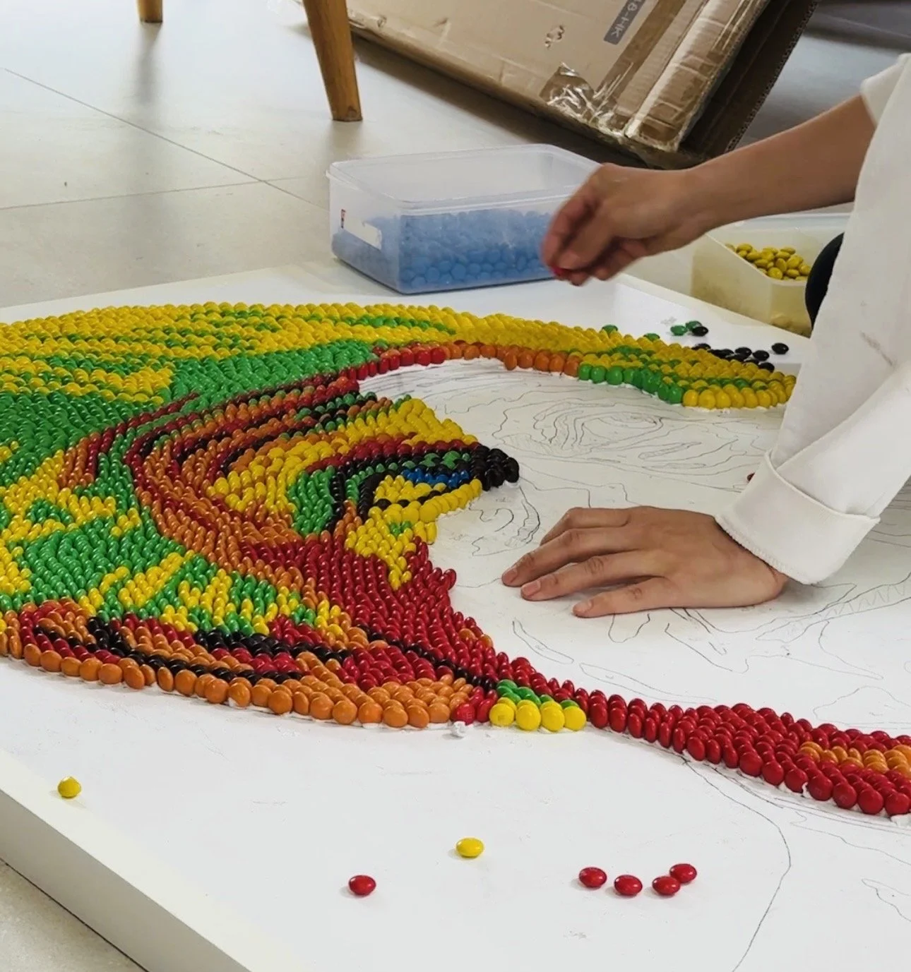 Close-up shot of food artist Michelle Wibowo's hands meticulously arranging red, yellow, and green M&M chocolates on a canvas to create a pop art mosaic portrait of King Charles III, following a detailed pencil sketch outline.