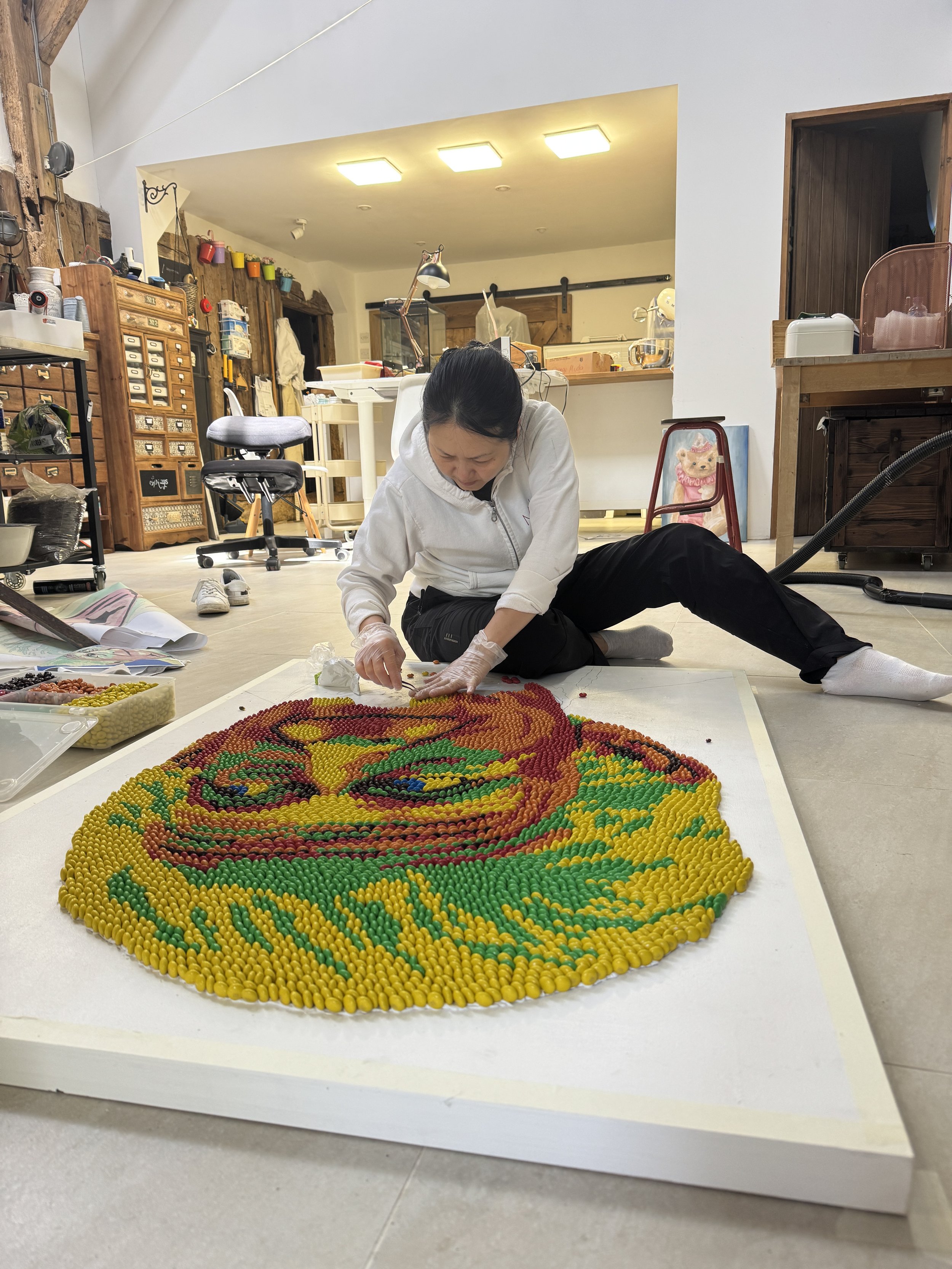 UK food artist Michelle Wibowo in her studio, sitting on the floor to meticulously assemble a pop art mosaic portrait of King Charles III using thousands of colorful M&M chocolates on a canvas.